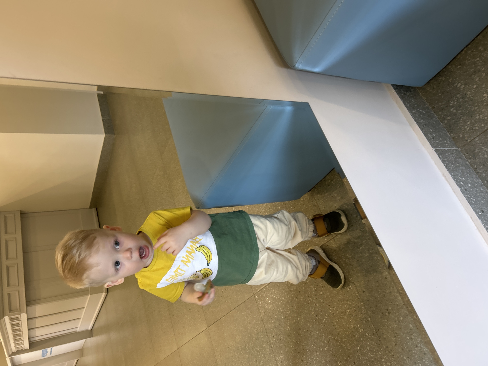A toddler is standing on a terrazzo floor next to a light blue bench in what appears to be a public space, possibly a museum or waiting area. The child has blond hair, is wearing a yellow t-shirt with a banana design, green bib overall, and beige pants with brown sandals. The walls are beige. The location cannot be determined to be in the city of Izhevsk, Russia based on the given image.