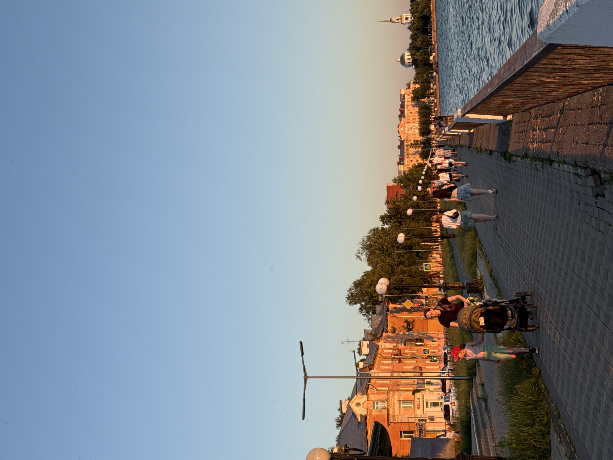 The image shows a waterfront promenade in Votkinsk, Russia, during a sunset. A wide paved walkway runs alongside a body of water, separated by a metal railing. On the left side of the walkway, there is a grassy area and buildings, including an orange-colored apartment building with multiple windows. Several lampposts line the promenade.
In the foreground, a woman is pushing a stroller with a child walking beside her. Further down the promenade, a group of people are strolling, walking in various directions. In the background, more buildings and trees are visible, and a church with a tall spire can be seen in the distance. The sky is clear and light blue, with a warm, golden hue cast by the setting sun. The water appears calm with gentle ripples.