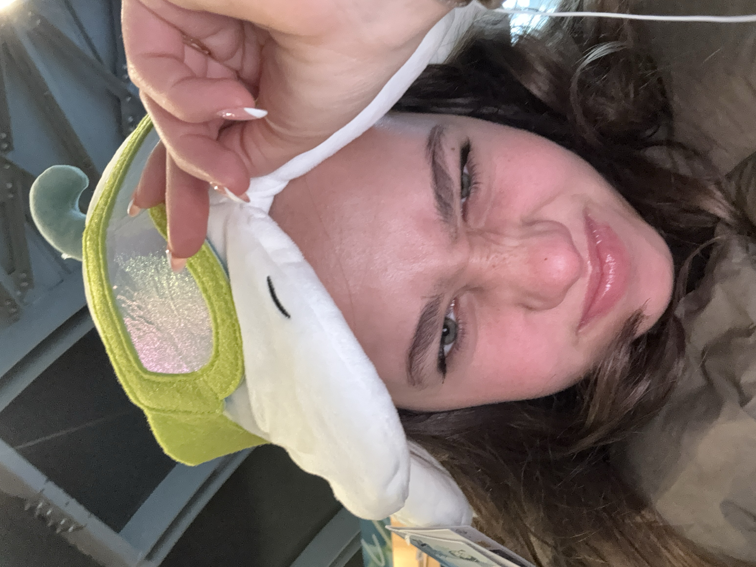 The image captures a close-up of a female individual located in the city of Rho, Italy. The subject, facing forward, has dark brown hair and olive green eyes, displaying a scrunched facial expression with narrowed eyes, furrowed eyebrows, and a slight smirk. She is wearing a white plush hat featuring a prominent lime-green, reflective visor-like brim and a small, light blue curved appendage at the top. Her left hand, adorned with an almond-shaped nail exhibiting a white French tip, is visible pinching the white fabric of the hat on the top-right side. A white earbud wire descends along the right side of her head. The background is out of focus, showing structural elements of a ceiling or large beam, characterized by grey-blue tones and visible rivets, consistent with an indoor commercial or public venue.