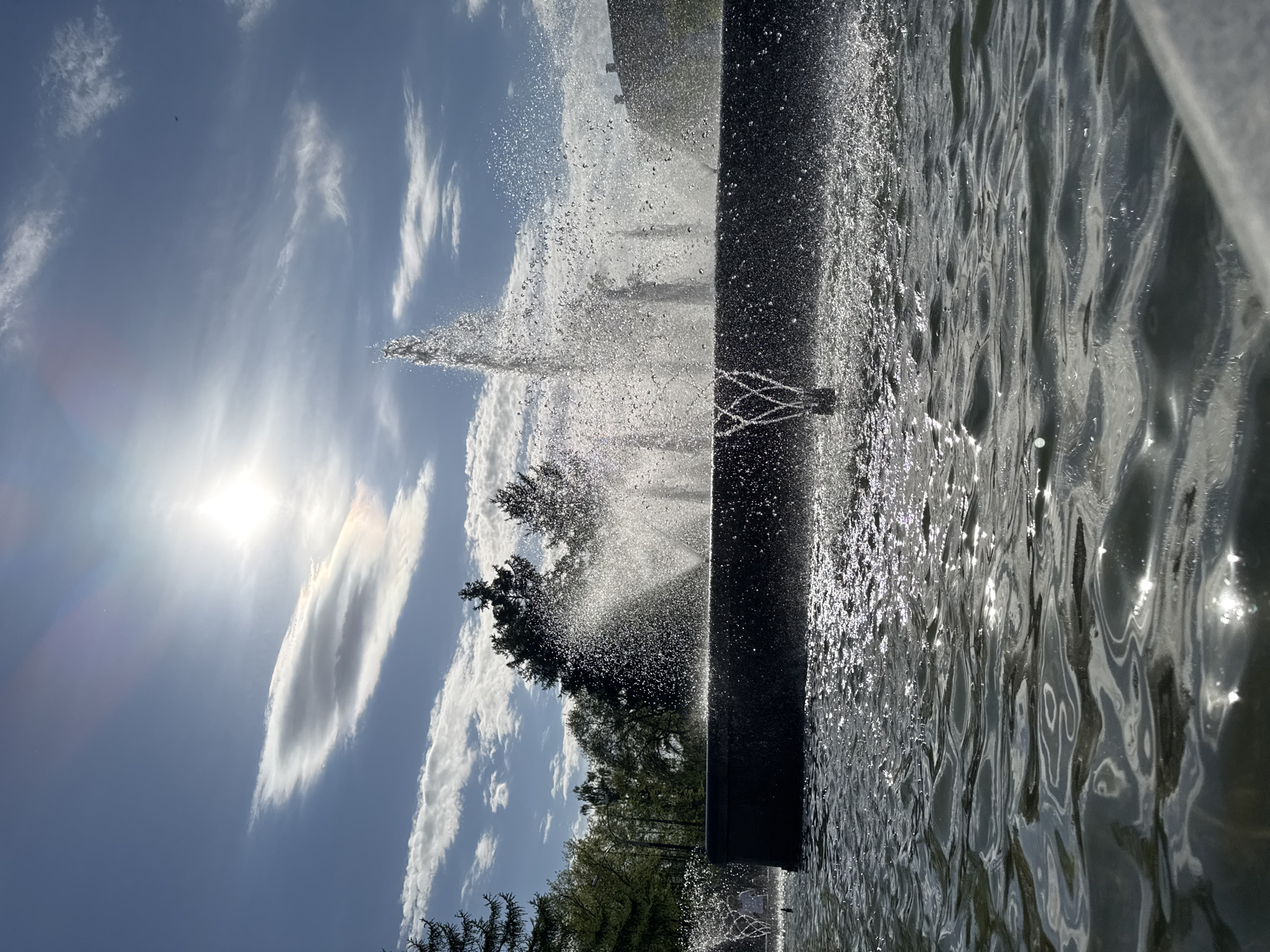 The image shows a fountain in the city of Chaikovsky, Russia. Water is being propelled into the air, with the sunlight creating reflections and sparkles. The sky is blue with streaks of clouds. There are trees in the background. The fountain's structure is visible, with water cascading around it. The water in the pool appears rippled.