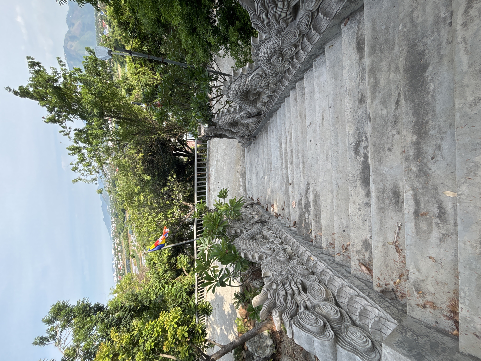 The image shows a downward view of a concrete staircase with ornate dragon carvings on the railings. The staircase is located outdoors amidst lush greenery and trees. In the background, a town or city with buildings and mountains can be seen under a bright, hazy sky. A flagpole with a colorful flag, likely representing Buddhism, is visible to the left of the stairs. The scene appears to be situated in Tay Nha Trang Ward, Vietnam, given the architectural style and landscape. There are no people visible in the image. The environment is somewhat weathered, with fallen leaves scattered on the stairs.