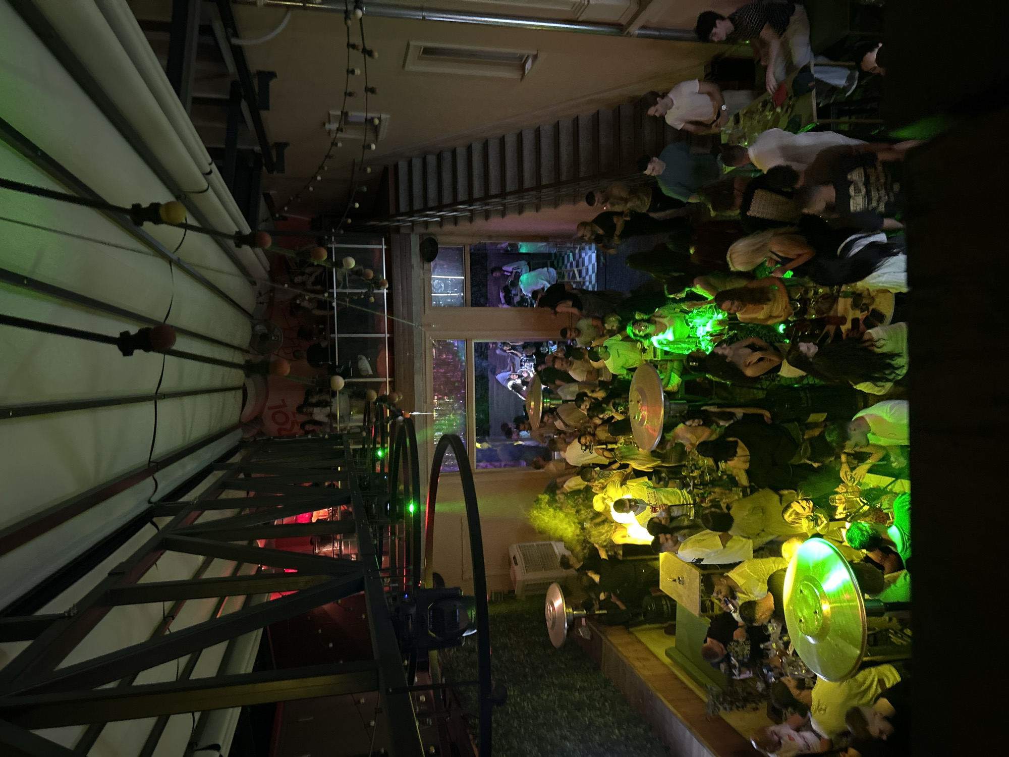 The image depicts an outdoor dining area in Plovdiv, Bulgaria, at night. People are seated at tables, illuminated by overhead lighting. A building with a staircase and window is in the background. Metal structures are visible above the seating area.