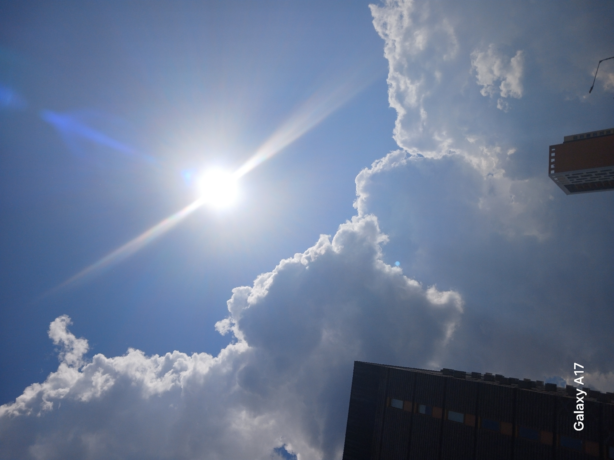 The image shows a daytime sky with the sun shining brightly, creating lens flares. White, puffy cumulus clouds fill the majority of the frame, contrasting against the blue sky. The lower portion of the image includes parts of buildings. A tall, dark-colored building with a textured facade is in the bottom right corner, and a section of a light brown building is in the upper right. The text "Galaxy A17" is printed vertically along the right edge of the image, suggesting the image was taken with a Samsung Galaxy A17 smartphone. The location, as inferred from the question, is Yekaterinburg, Russia. There are no people visible in the image. The dominant actions are the atmospheric processes creating clouds and the sun shining.