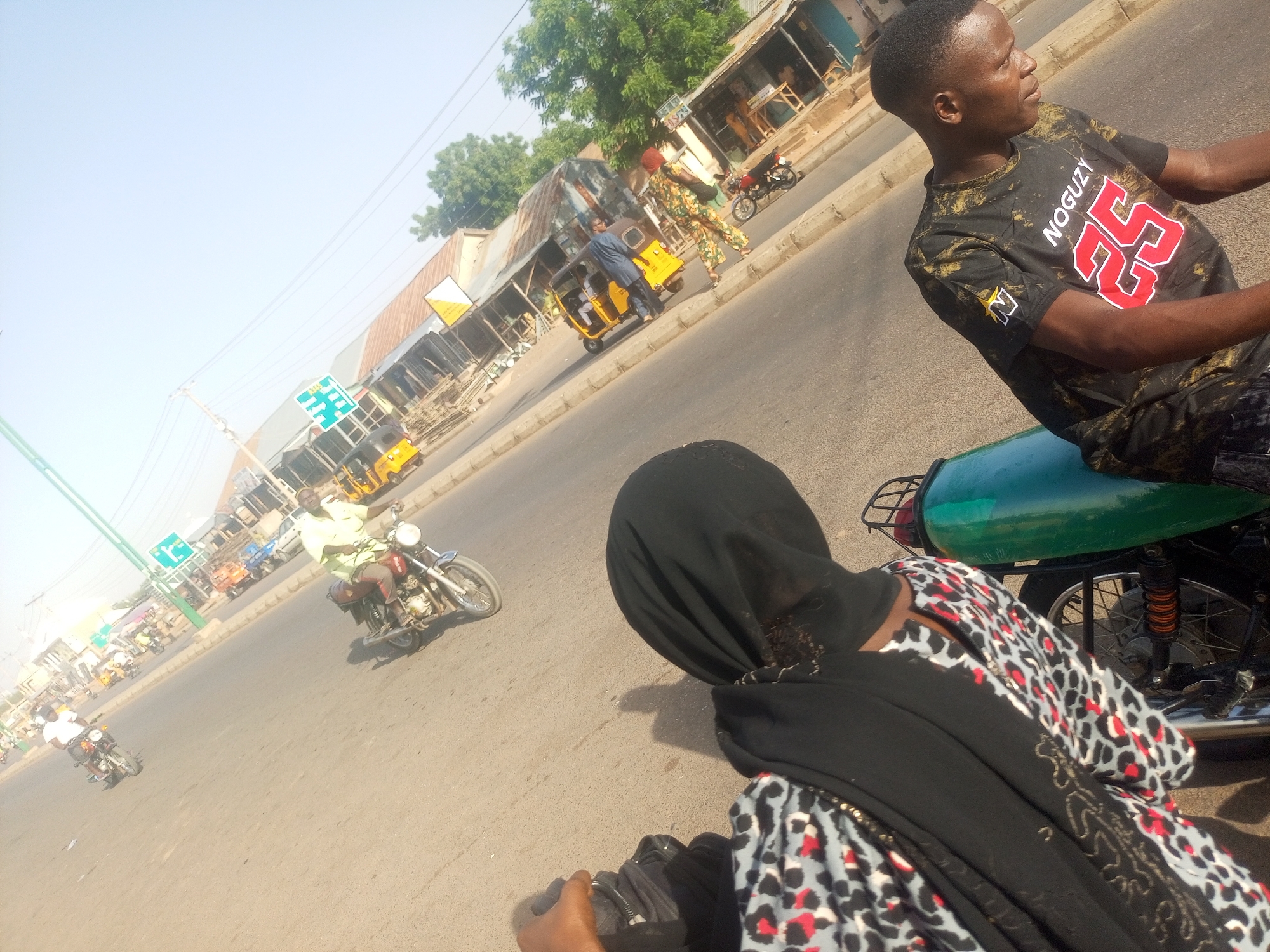 The image depicts a street scene in Tempure, Nigeria. A man wearing a black T-shirt with the word "NOGUZY" and the number "25" on it is riding a green motorcycle. Behind him sits a person wearing a black headscarf and a patterned top, possibly a dress.
In the background, there is a road with other vehicles, including motorcycles and yellow auto rickshaws. Buildings line the street with various shops and stalls. A traffic sign is visible in the distance. Other people are on the sidewalks. The environment appears to be a bustling commercial area in Tempure.