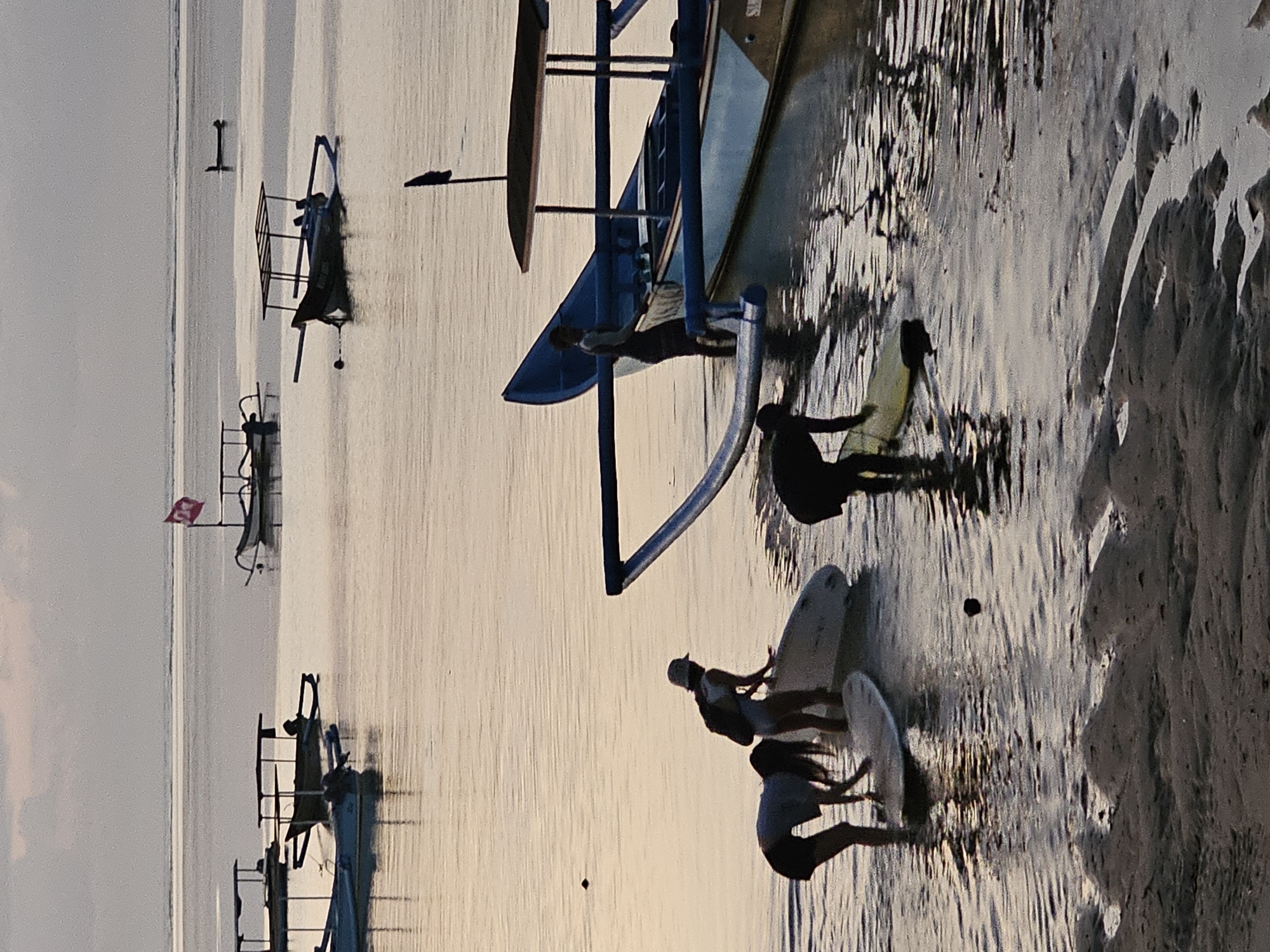 In Denpasar, Indonesia, at dawn, a group of people are preparing to surf in the calm, shallow waters of the ocean. The image captures several individuals wading in the water, some with surfboards. One person is bending down to adjust their surfboard, while another stands near a larger boat. Several traditional fishing boats, known as jukungs, are anchored in the serene water, some with thatched roofs and outriggers. In the background, a few more jukungs are visible, one adorned with a red flag. The sky is overcast with soft, diffused light, indicating the early morning hour. The foreground shows a sandy beach with rippled patterns from the receding tide. The overall scene depicts a tranquil start to a day of surfing and fishing.