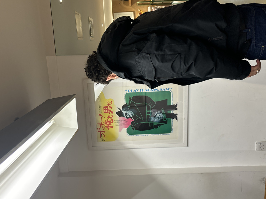 A lone individual is depicted indoors, viewed from a rear-right perspective, observing a framed poster. The subject, an adult male with dark, curly hair, wears a black, multi-pocketed jacket, dark blue jeans, and a silver-colored ring on his right hand's ring finger. He stands facing a white wall on which a framed Japanese film poster is mounted. The poster, predominantly yellow at the top and green at the bottom, features a stylized caricature of Humphrey Bogart in a trench coat and fedora, smoking, with pink smoke emanating. The Japanese title "ボギー!俺も男だ" (Bogie! I'm a Man Too) is prominent at the top, while the English title "PLAY IT AGAIN, SAM" runs vertically along the right side. Two smaller portraits, one male and one female, with "CASABLANCA" partially visible on the female's, are incorporated into the Bogart figure. Small, largely illegible English text is present vertically along the left side. A white fluorescent light fixture with a grey casing illuminates the scene from the ceiling directly above. A white Type G electrical outlet is affixed to the lower left portion of the wall. The blurred background to the right suggests an open indoor space. Based on the Type G electrical outlet, this scene is located in an indoor establishment within London, United Kingdom.