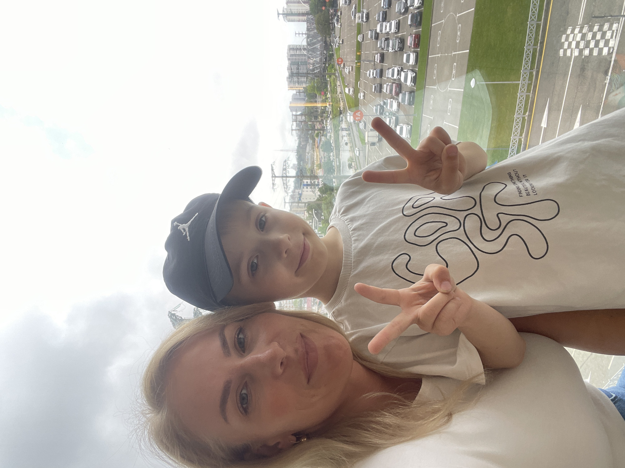 A woman and a young boy are taking a selfie from a high vantage point in Sochi, Russia. The woman is at the bottom of the frame with the boy above her, both giving the peace sign. The boy is wearing a grey baseball cap with a white logo. Behind them, visible through a window, is a view of the city including buildings, roads, and a parking lot filled with miniature cars. The environment is overcast.