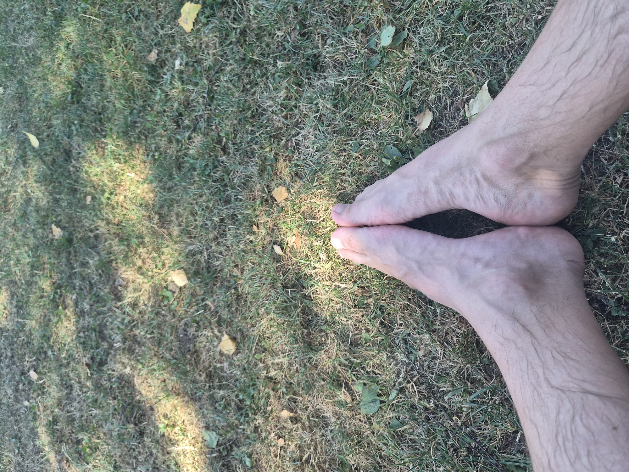 The image shows a pair of bare human feet positioned in a V-shape on a patch of grass. The grass is a mixture of green and dry blades, with a few fallen leaves scattered across it. Sunlight casts dappled shadows on the ground. The feet appear to be a light skin tone, with visible veins and fine hairs on the lower legs. The toes of both feet are visible and are positioned close together. The background consists entirely of the grassy ground. There are no indications of a city environment or specific landmarks that would allow for pinpointing a location within Landl, Austria. The image is a close-up shot focused solely on the feet and the immediate ground.