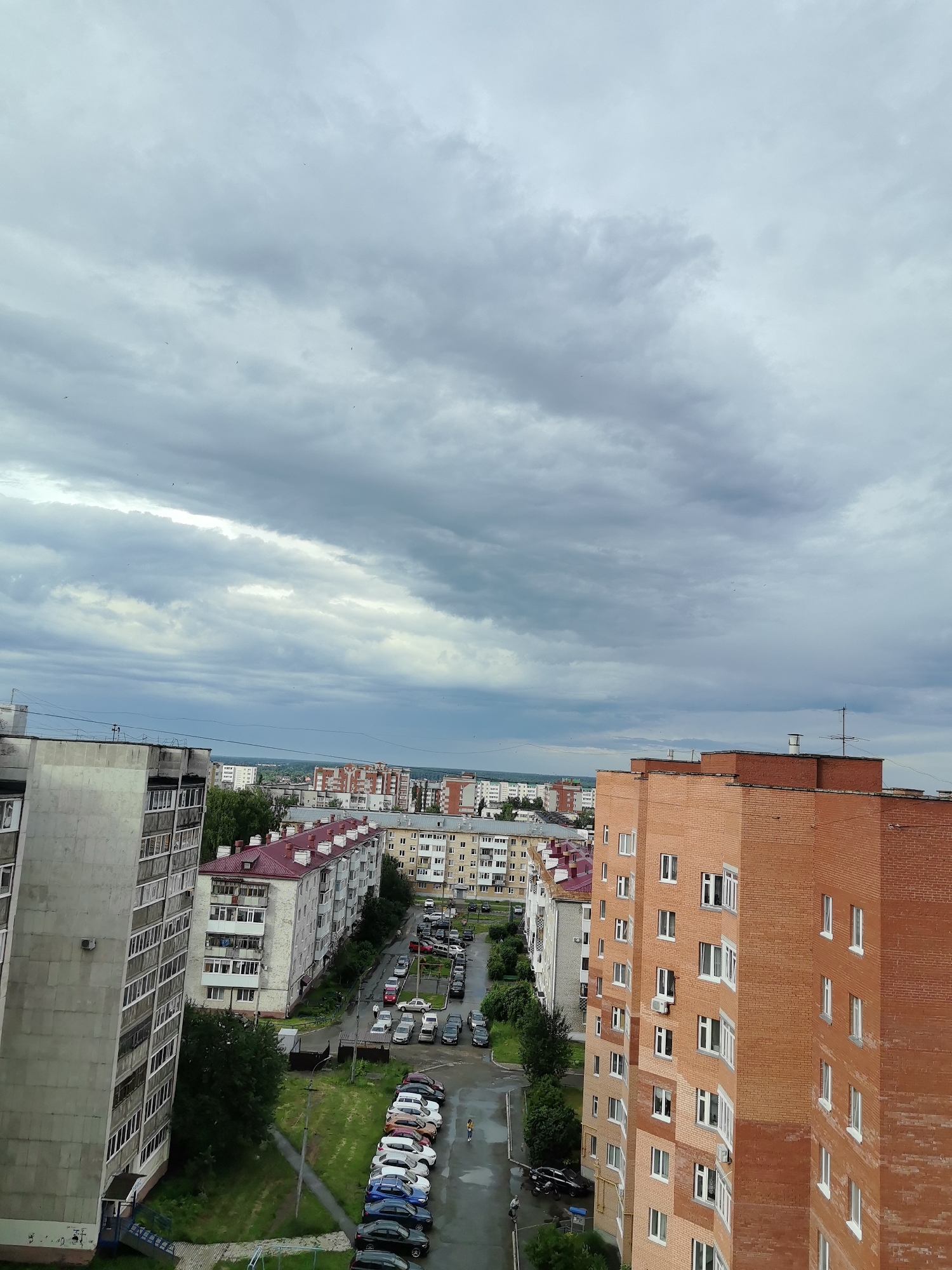 The image is an aerial view of a city street in Neftekamsk, Russia, under a cloudy sky. On the left, a tall, grey apartment building dominates the foreground. To its right, a row of apartment buildings with red roofs stretches into the distance. Further into the scene, a bright orange brick apartment building stands prominently on the right side of the image. The street below is lined with parked cars, and there are patches of green grass and trees between the buildings and the street. A few pedestrians are visible on the street and sidewalks. The sky is overcast with heavy grey clouds, with some lighter patches suggesting a break in the cloud cover. The overall impression is a typical urban residential area in a moderately developed city.