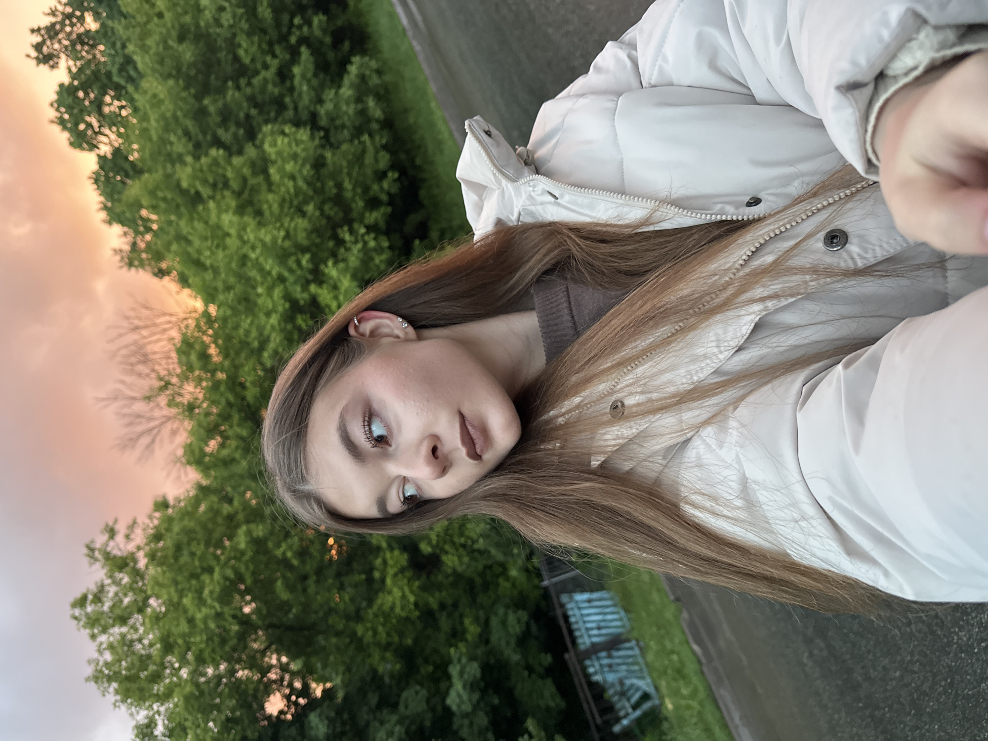 The image is a portrait of a young woman taken outdoors, likely a selfie, with the original orientation rotated 90 degrees counter-clockwise. The woman has long, light brown hair flowing over her shoulders and down her chest. She is wearing a light beige or off-white puffer jacket over a brown top. Her left ear is visible, adorned with two small, silver-toned hoop earrings, one featuring a tiny gem. Her face shows subtle makeup, including defined eyebrows and neutral-toned lipstick, and she gazes calmly to her left (viewer's right) with a slightly pensive expression.
The background depicts a natural outdoor setting during the "golden hour," characterized by a sky displaying warm hues of orange and pink, suggesting either sunrise or sunset. Below the horizon, a dense expanse of lush green trees fills the middle ground, indicating a park or wooded area. The foreground and lower background feature a dark, paved surface, possibly a road or pathway, alongside a patch of green grass. A white or light-colored fence or railing is faintly visible in the distance on the ground level. There is no visible text in the image. The scene is set in Kazan, Russia.