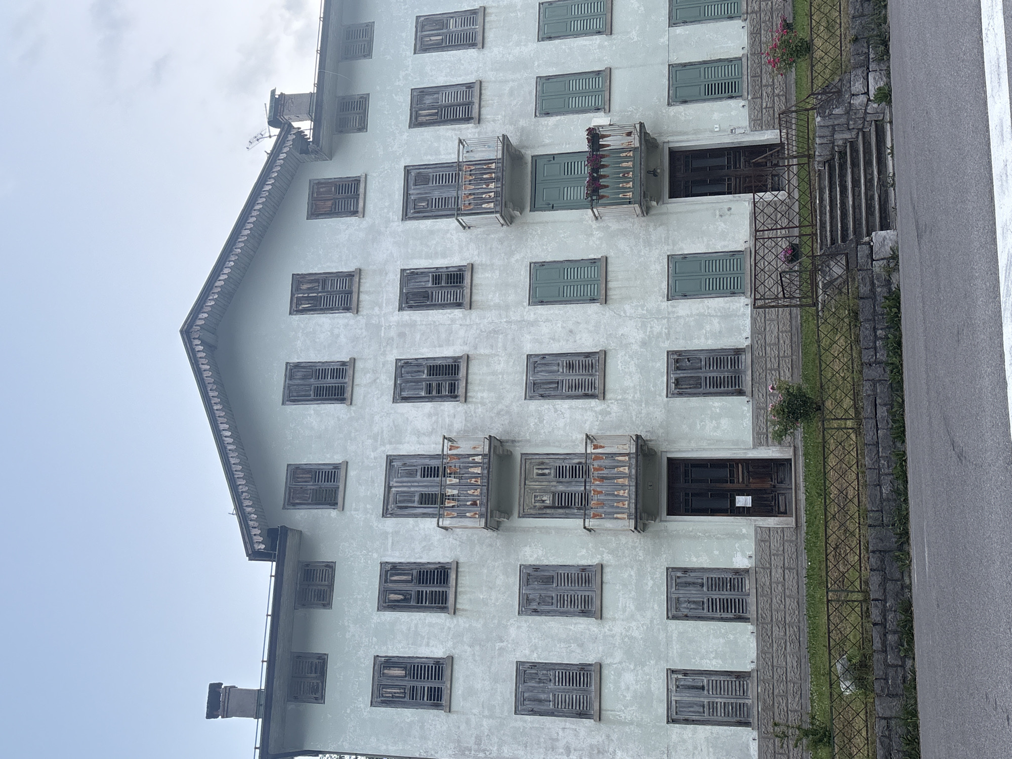 The image shows a tall, multi-story building with a light green, textured facade. The building has a steeply pitched roof with decorative trim and a chimney on the left side. The facade is covered in numerous windows, many of which have weathered wooden shutters. Some of the shutters are partially open, revealing darker interiors. There are also several balconies with decorative iron railings.
In the foreground on the right side of the image is a paved road. To the left of the road, there is a stone staircase leading up to a dark wooden double door on the building. The staircase is flanked by a metal fence and bordered by grass and some flowering plants. The overall impression of the building is one of age and perhaps disuse, with peeling paint and weathered materials.
The sky is overcast and appears bright, suggesting it is daytime. The lighting is diffused, and there are no strong shadows, indicating a cloudy or misty day. The scene appears to be in a somewhat rural or mountainous setting, given the architecture and the presence of natural elements. There is no visible text in the image.