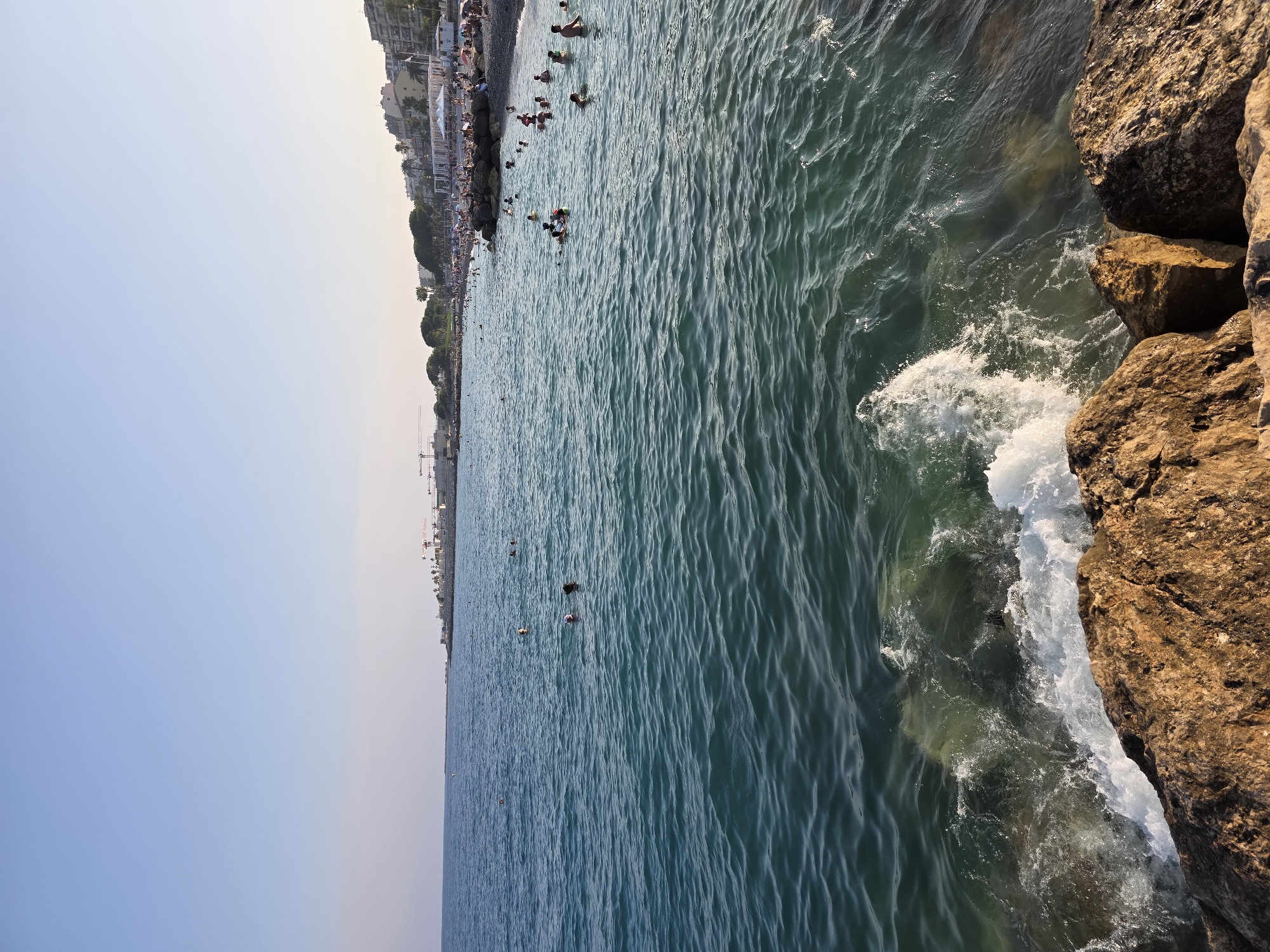 The image depicts a view of the Mediterranean Sea at the Promenade des Anglais in Nice, France, on a clear, sunny day with a pale blue sky. The foreground is dominated by large, weathered rocks, some of which are wet from the sea spray. Waves are breaking against these rocks, creating white foam. The sea itself is a deep teal color, with ripples and small waves visible.
In the mid-ground and background, numerous people are enjoying the water, swimming and wading. Some are closer to the shore, while others are further out. Along the coastline, a populated beach is visible, lined with buildings that appear to be hotels and apartments. Palm trees and other greenery are interspersed with the structures. Several construction cranes are visible in the distance, suggesting ongoing development. The scene is bustling with activity, characteristic of a popular tourist destination.