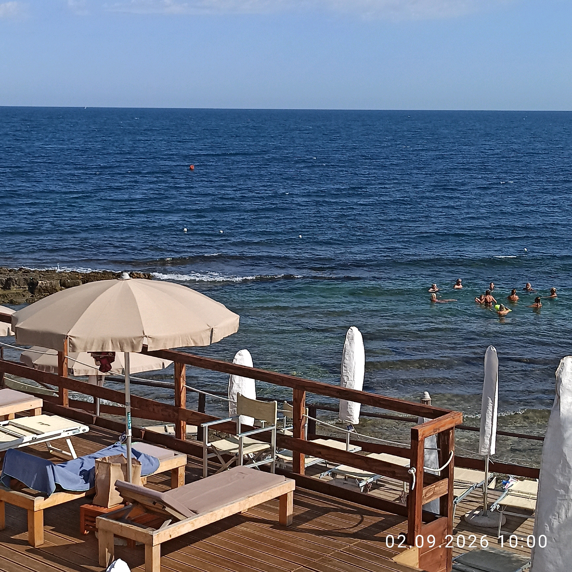 The image captures a sun-drenched scene at a waterfront location in Molfetta, Italy, likely a beach or a lido. In the foreground, a wooden deck is furnished with several lounge chairs, some occupied by blue and beige towels and a brown tote bag. A large beige umbrella provides shade over one of the lounge areas. A wooden railing separates the deck from the sea.
Beyond the deck, the tranquil blue waters of the sea stretch out to the horizon under a clear sky. Scattered across the water are several people, some appearing to be swimming or wading, enjoying the water. The water near the shore is clear, revealing rocky formations beneath the surface.
On the deck, two tall, white, cylindrical objects, possibly covered beach umbrellas or decorative elements, stand upright. The scene is dated "02.09.2026 10:00," indicating it was captured in the morning on September 2nd, 2026. The overall atmosphere is one of leisure and relaxation in a coastal setting.