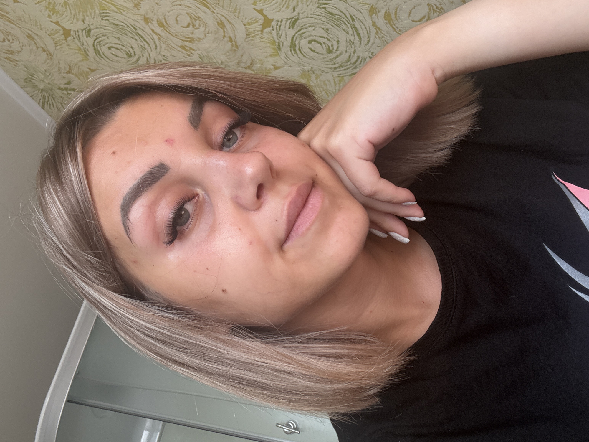 A young woman with blonde, shoulder-length hair is shown in a close-up, shoulder-up portrait. She is wearing a black t-shirt and is resting her chin on her hand. Her makeup includes false eyelashes and defined eyebrows. There is a red blemish on her forehead and some other minor skin imperfections. The background consists of a textured, light green and yellow wallpaper with a swirling pattern. To the left, a sliver of a modern shower enclosure is visible. The overall lighting suggests natural daylight. The scene is indoors, likely in a residential setting in Krasnogorskoye, Russia.