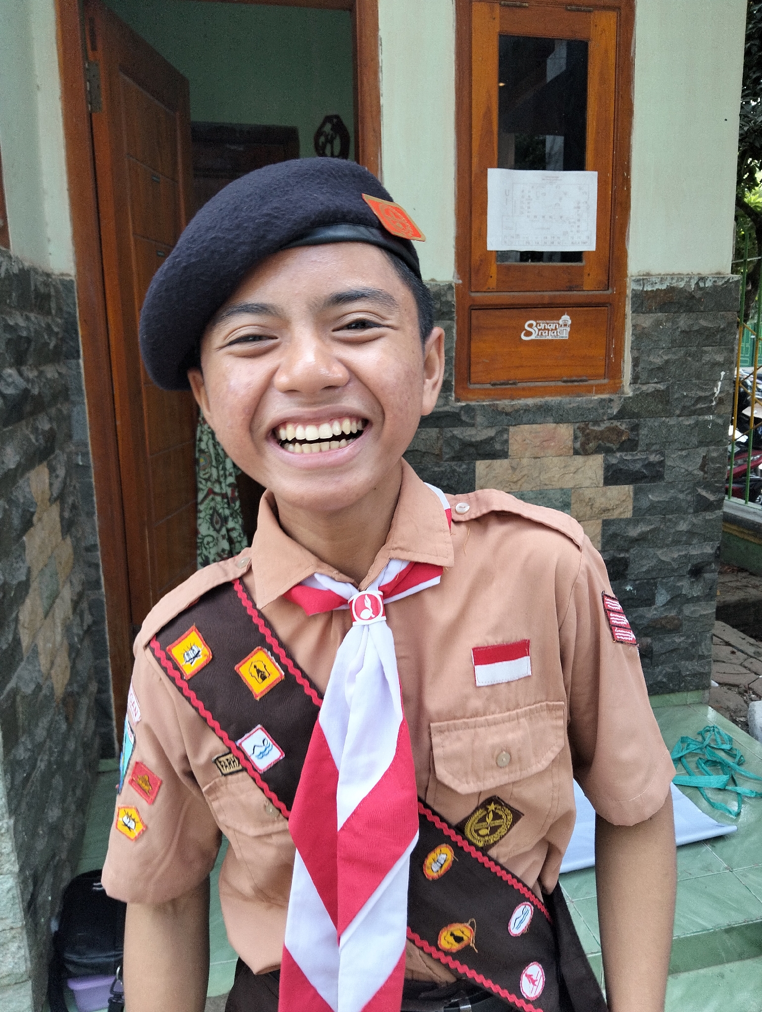 A boy wearing a scout uniform with a black beret and a red and white scarf is smiling at the camera. He is standing in front of a wooden door and a stone wall. The scout uniform has several badges and patches. A white piece of paper is taped to the door, and a logo that reads "Sunanrajat" is visible on the door. The background appears to be an outdoor setting with some greenery.