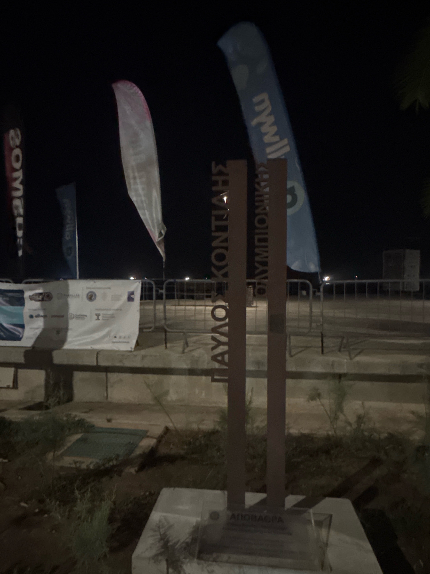 The image shows a nighttime scene in Limassol, Cyprus, featuring a metal monument with Greek text. The monument appears to be located outdoors, near a promenade or waterfront, as indicated by a metal barrier and a white banner with logos in the background. The text on the monument reads "ΠΑΥΛΟΣ ΚΟΝΤΙΑΔΗΣ ΟΛΥΜΠΙΟΝΙΚΗΣ" and below that "ΑΠΟΒΑΣΗ". Surrounding the monument, there is sparse vegetation and a concrete base. In the background, several flags and banners are visible, some with text. The overall lighting suggests it is dark, with artificial lighting present.