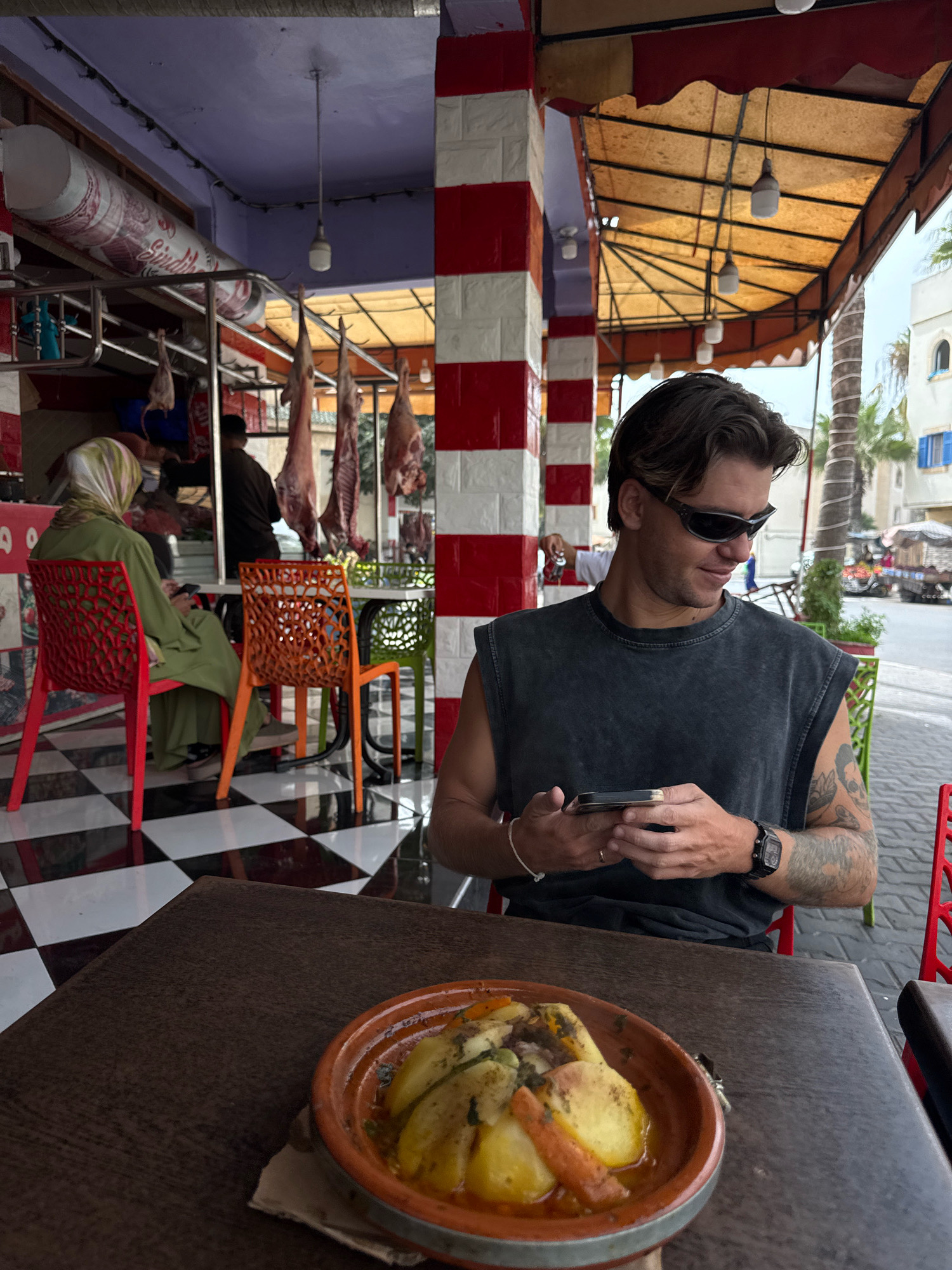 The image shows a man in sunglasses and a dark sleeveless shirt sitting at a table with a dish of food. He is holding a smartphone in his hands. Behind him, there is a butcher shop with meat hanging from the ceiling and a woman wearing a headscarf sitting on a red chair. The floor has a black and white checkered pattern. The scene appears to be in Essaouira, Morocco, as suggested by the architecture and the presence of a tagine dish, a traditional Moroccan stew. The overall atmosphere is casual, with elements of a marketplace or a roadside eatery.