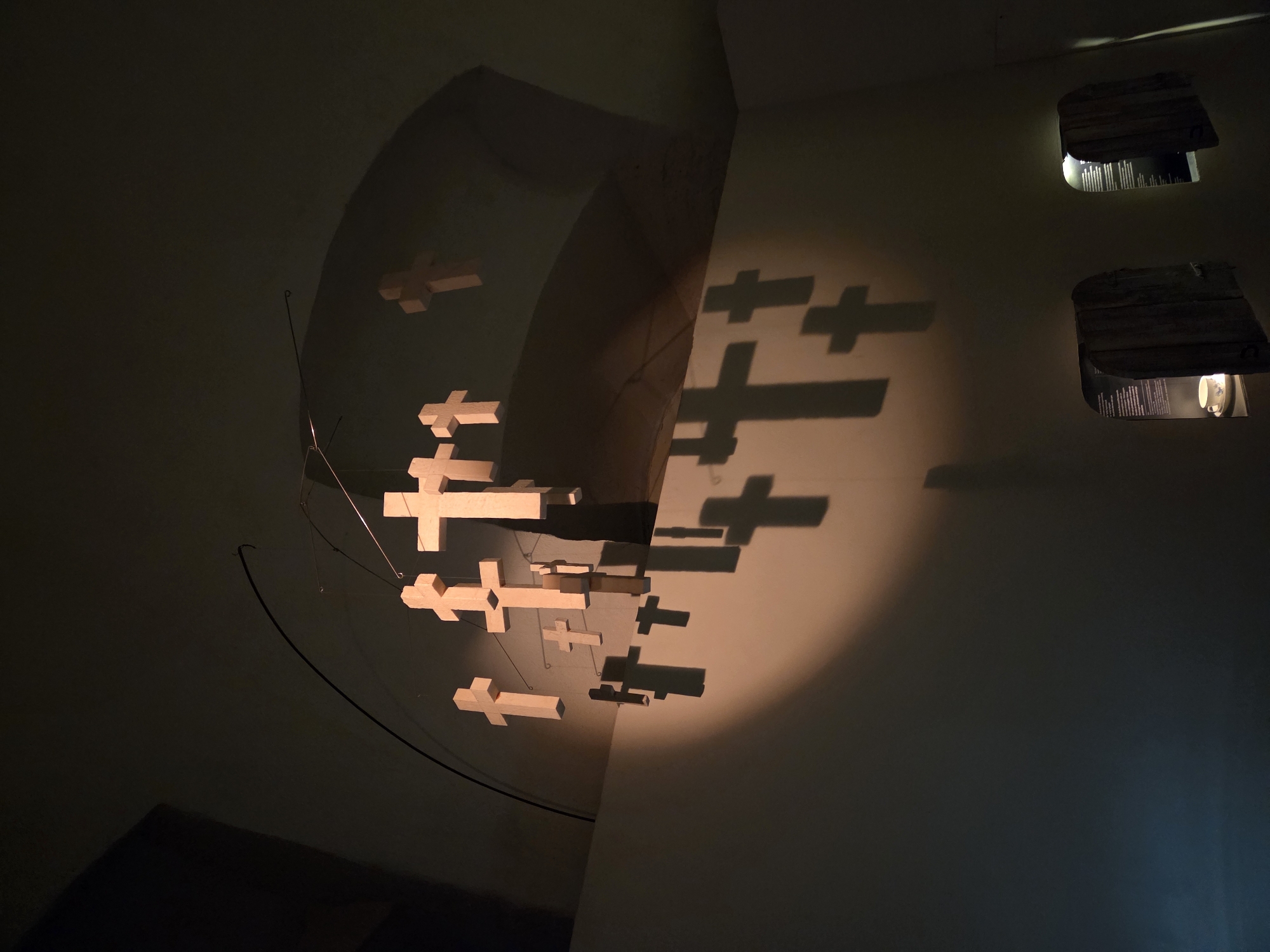 The image shows a mobile of wooden crosses suspended from a thin wire that curves across a dark, arched ceiling. The crosses are illuminated by a spotlight, casting large shadows on a light-colored wall. The mobile is comprised of multiple crosses of varying sizes, some hanging closer to the wall than others. The overall scene is dimly lit, with the spotlight on the crosses being the primary light source. The environment appears to be an indoor space, possibly a museum or exhibition hall, with rough-textured wooden structures visible at the bottom right of the image, which might be display cases. The location is specified as Malmö, Sweden.