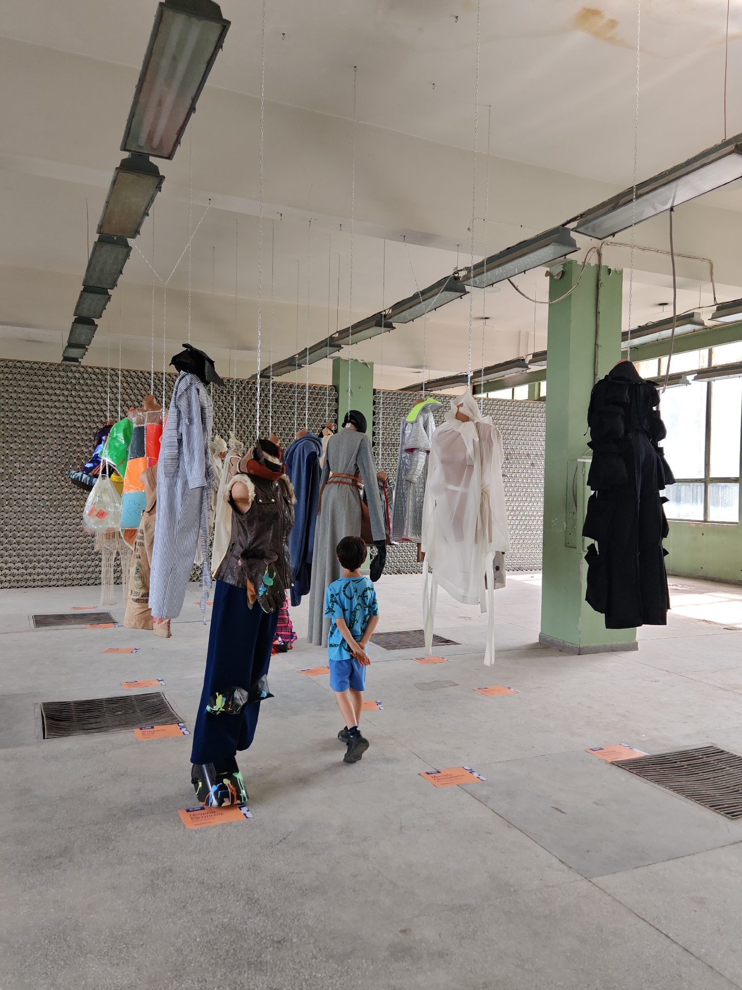 This image depicts an indoor exhibition space, presumed to be in Gabrovo, Bulgaria, characterized by a raw industrial aesthetic. The high, off-white ceiling is fitted with a series of long, rectangular fluorescent light fixtures, some exhibiting minor rust-colored staining. Numerous thin metal chains descend from the ceiling, suspending various garments and fashion objects as part of an installation.
On the left side, a patterned wall constructed from stacked, grey-toned cylindrical elements forms the background. Towards the right, a prominent square-sectioned support column, painted light green, stands. Beyond this column, large multi-paned windows allow natural light to illuminate the space. The floor is grey concrete, featuring several embedded rectangular metal grates. Orange square informational markers are placed on the floor; visible text includes "Wikipedia" and "Elena Kirova" on one, and "Manuela Georgieva" on another.
The suspended exhibits, arranged across the scene, include: a cluster of colorful plastic bags (orange, green, clear, blue, red) alongside a beige burlap sack with partially legible text (e.g., "JUMBO"); a long, vertically striped white and light blue shirt topped with a dark hat; a torso-like figure dressed in a brown leather-style vest with fur detailing, dark blue trousers, and shoes splattered with multi-colored paint; a figure wearing a long grey coat or dress cinched with a brown belt and a dark hat; a translucent white garment, possibly a sheer coat, suspended on a white framework; and a voluminous, layered black garment hanging near the green column.
A single individual, a young boy estimated to be between five and eight years old, is present in the middle ground, his back mostly turned to the viewer. He wears a blue short-sleeved t-shirt with a lighter blue pattern and light blue shorts. His hands are clasped behind his back as he looks upwards and to the right, observing the suspended exhibits.