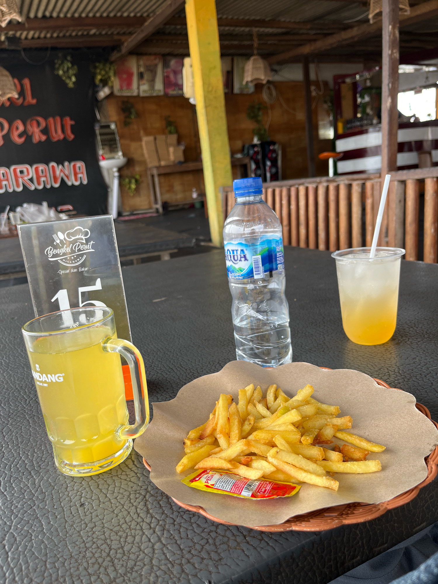 An outdoor table at a restaurant in Tuntang, Indonesia, is set with a meal and drinks. On the table is a plate of french fries with a packet of chili sauce, a glass mug filled with a yellow beverage and a straw, a clear plastic cup with a yellow drink and ice, and a bottle of Aqua brand water. A table tent with "Bengkel Perut" and the number "15" is also visible. In the background, wooden structures and railings suggest an open-air dining area.