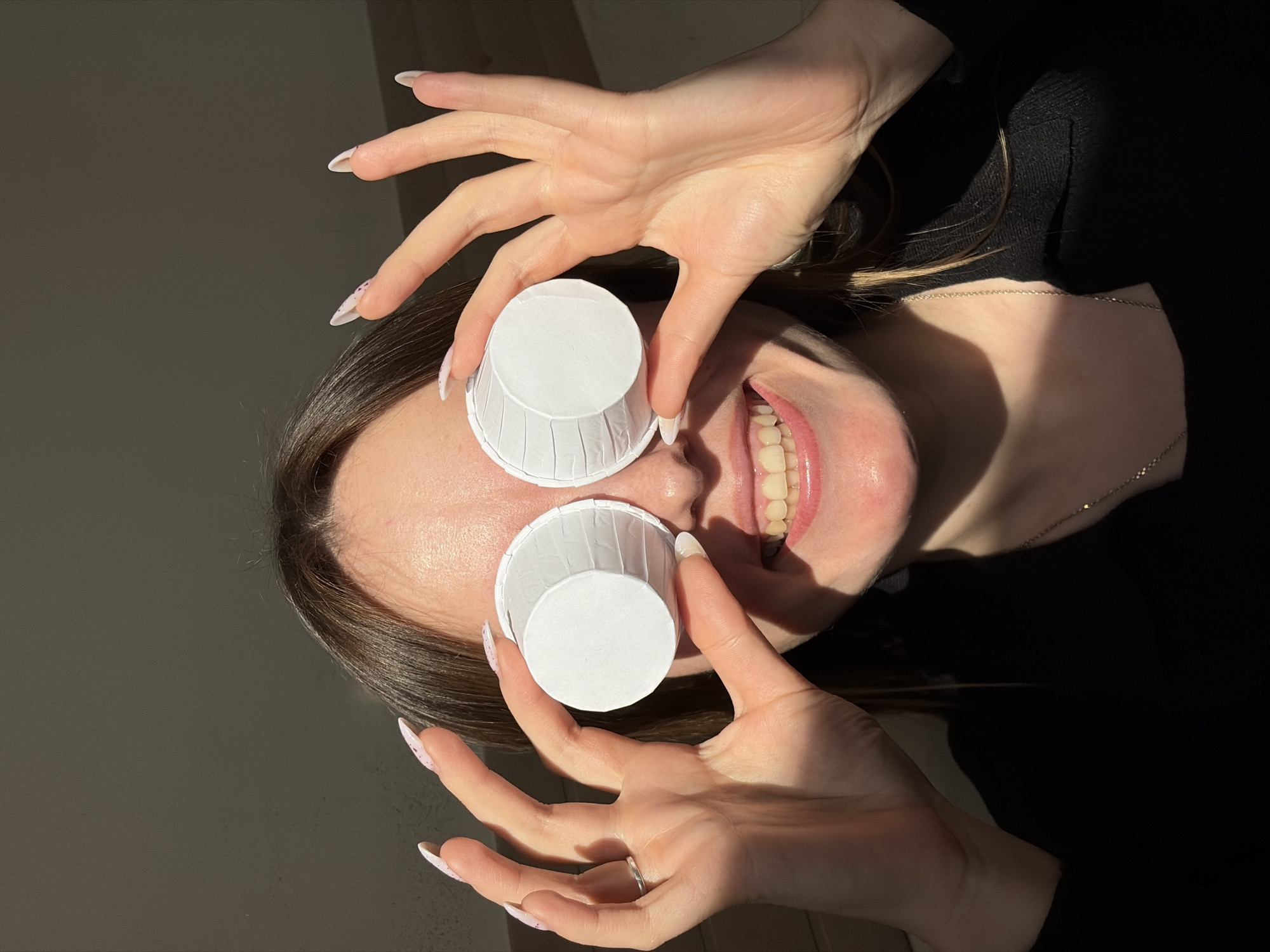 A young adult female is holding two small white paper cups over her eyes, mimicking wearing glasses. She is smiling with her teeth visible. Her hair is dark brown, and she is wearing a black top. Her fingernails are long and painted with a light color and a purple speckle design. She is wearing a necklace. The background appears to be a wall in a room with sunlight casting shadows. The location is unknown.