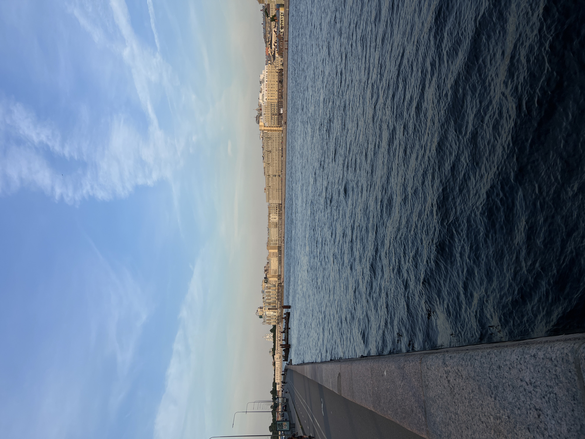 The image shows a wide river in Saint Petersburg, Russia, with the city's embankment and buildings in the background. The river is dark blue and choppy, with small waves. On the left side of the image, there is a granite embankment, a paved walkway, and a bike path. Streetlights line the path, and there are a few people on bicycles and walking. In the distance, across the river, is a long line of buildings with various architectural styles, including classical and modern. The sky is blue with wispy white clouds, suggesting it is either early morning or late afternoon. The overall impression is a peaceful urban landscape.
