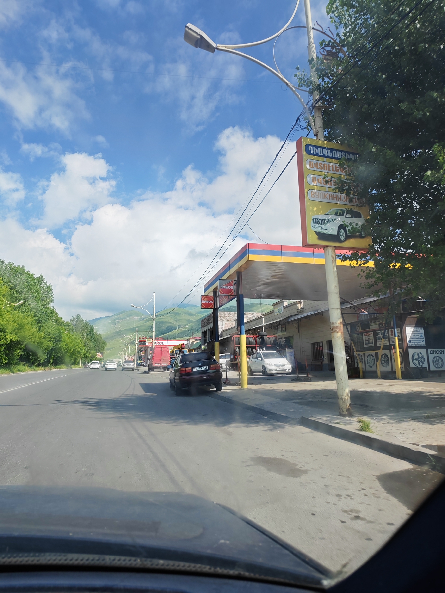 The image captures a scene along a road in Vanadzor, Armenia, likely on a sunny day given the bright lighting and scattered clouds. The perspective is from inside a vehicle, looking forward.
The foreground shows the dashboard and windshield of the car, indicating the photographer is a driver. The road itself is paved and appears to be a multi-lane highway. Several cars are visible on the road, some driving ahead and one parked on the right side.
To the right of the road, there is a gas station with a brightly colored canopy (red, yellow, and blue). The station appears to be branded with "ENEOS." Adjacent to the gas station is a building that seems to house a tire shop, with stacks of tires visible. A white car is parked in front of this shop. A large yellow sign with text in Armenian and what appears to be Cyrillic is mounted on a pole near the gas station, depicting a white SUV and offering services like tire repair ("ВУЛКАНИЗАЦИЯ").
On the left side of the road, there is a lush green treeline and a gently sloping hill in the background, possibly part of the landscape surrounding Vanadzor. The sky is blue with distinct white clouds. A street lamp is visible in the upper left corner of the image.
The overall impression is a typical roadside scene with commercial establishments in a city or its outskirts. The presence of the gas station and tire shop suggests a location where travelers might stop for services. The license plate on the car parked on the right side reads "37 KF 543," which is consistent with Armenian license plate formatting.