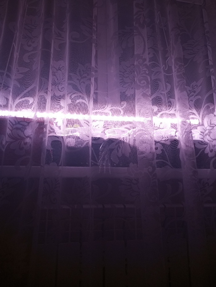 An indoor scene within the city of Яромаска, Russia, is depicted. The primary focus is a window covered by sheer, white or light-colored lace curtains, patterned with floral and scrollwork designs. A prominent, horizontal band of bright pink-purple light originates from behind the curtains, illuminating them from the approximate midpoint of the window. Through the semi-transparent fabric, the window frame and indistinct objects on the windowsill, possibly including a plant or container, are faintly visible. Below the window, in the darker lower portion of the image, a dark, grid-like structure, consistent with a radiator, is discernable through the curtains. No individuals are present. The environment suggests an interior space during low-light conditions, with the artificial light source serving as the main illumination.