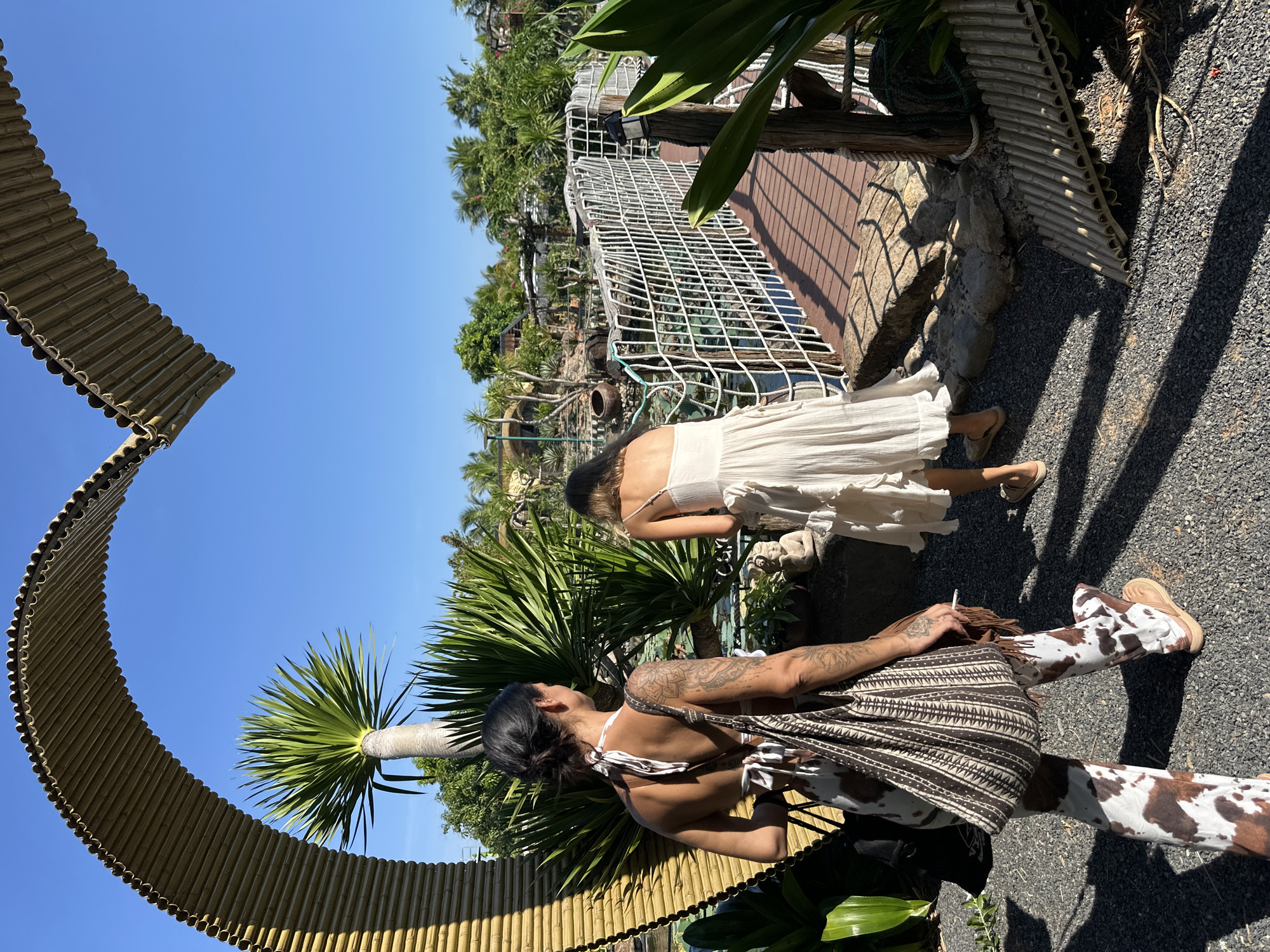 Two women are walking on a gravel path towards a wooden bridge in Mũi Né, Vietnam. The woman in the foreground, on the left, has dark hair pulled back and is wearing a white halter top with a brown and white patterned skirt and matching cow-print pants. She is carrying a large tote bag with a similar pattern over her left shoulder and has tattoos on her arms and back. The woman in the background, on the right, has blonde hair and is wearing a white ruffled dress. She is walking towards the bridge. The bridge has wooden planks and is bordered by a white rope fence. The environment is lush and tropical, with palm trees and other dense vegetation visible. A large, curved archway made of corrugated material, resembling a heart shape, is situated on the left side of the image. The sky is clear and blue, indicating a sunny day.