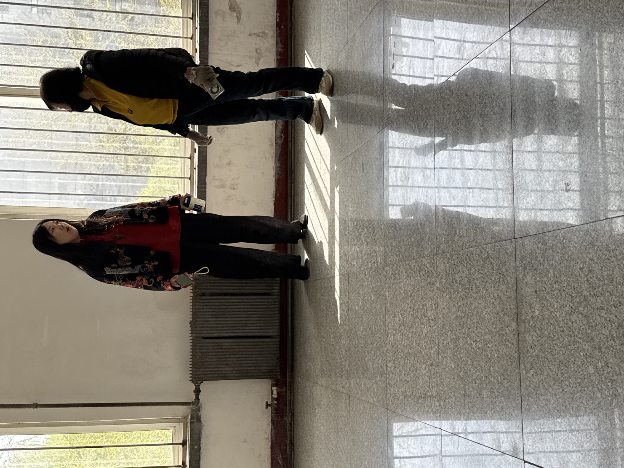 Two individuals are observed in an indoor location. The first person, on the left of the image, wears a red and black patterned jacket, black pants, and is holding a phone. The second person, on the right, wears a black jacket over a yellow shirt, jeans, gloves, and a black face mask. They are holding a small white item. They are standing by the window in the location. The floor is composed of square tiles. The environment contains windows, shadows of a gridded structure cast on the floor, and a radiator. The location is likely in Zhongtun, China, based on the location information provided.