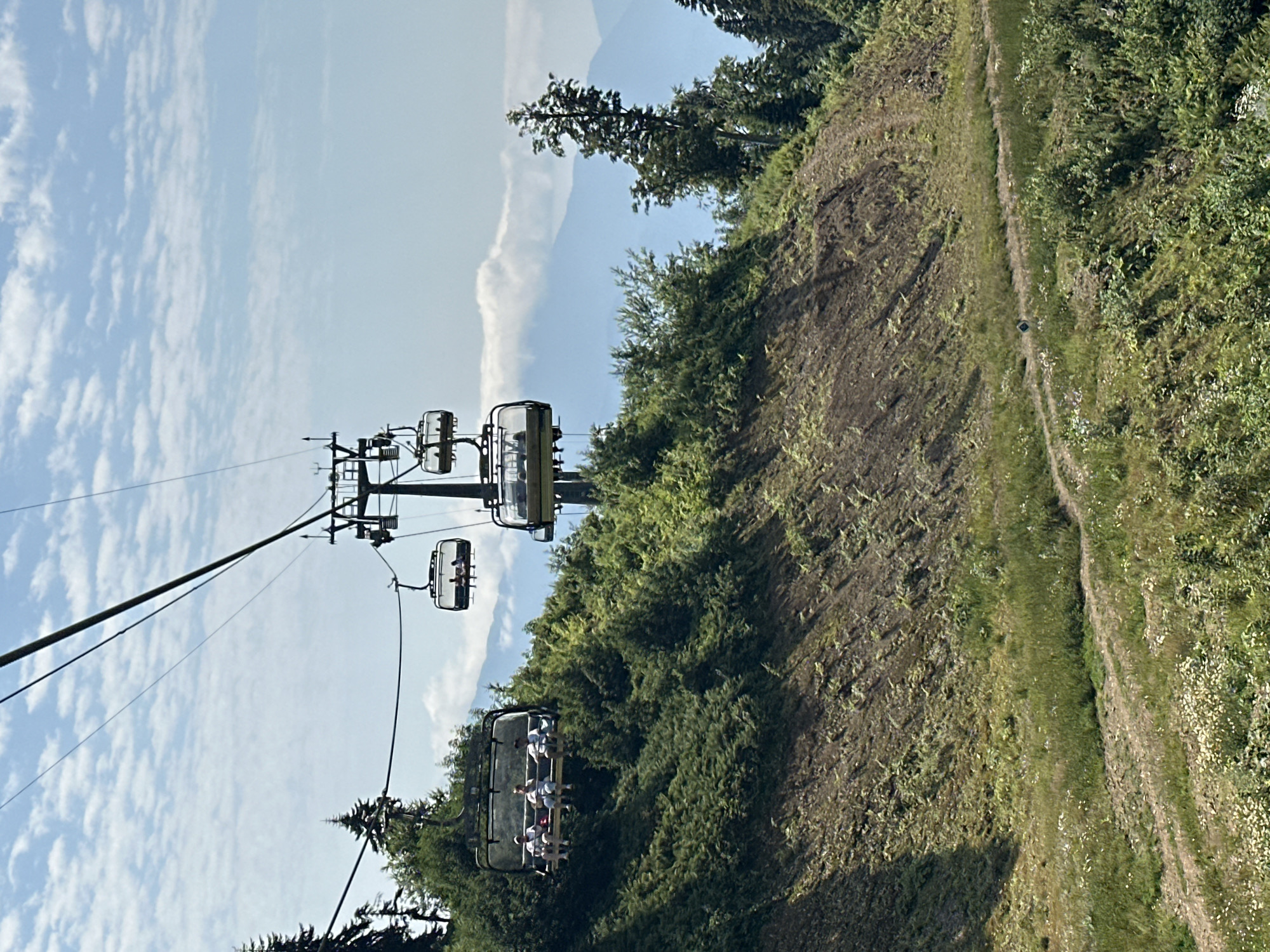 This image captures a scenic view of a ski lift operating on a lush green mountainside in Estosadok, Russia. The ski lift, with its dark metal structure and transparent gondolas, ascends against a backdrop of a bright blue sky dotted with white, wispy clouds. Several gondolas are visible, some carrying passengers. In one gondola, three individuals are seated, appearing relaxed as they enjoy the ride. The surrounding environment is characterized by dense vegetation, with the mountainside covered in a variety of green trees and shrubs. A winding dirt path is visible on the lower portion of the slope, adding a sense of depth and suggesting recreational opportunities in the area. In the distance, layers of blue mountains are faintly visible, hinting at the grand scale of the natural landscape. The lighting suggests it is daytime, with the sun casting shadows on the terrain.