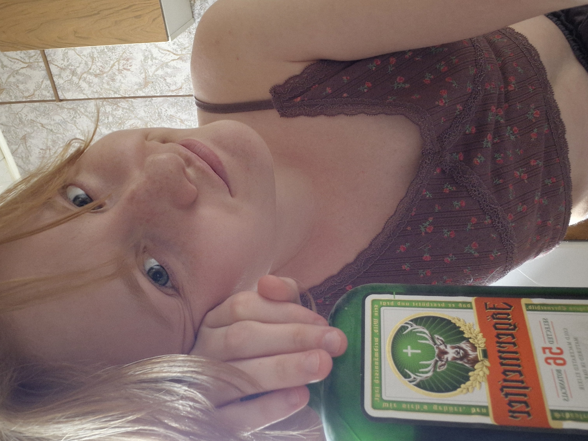 A fair-skinned person with red hair is lying on their side, holding a green bottle of Jägermeister. They have blue eyes and are wearing a dark brown floral-patterned camisole. The background includes what appears to be a tiled floor and a wooden surface. The image was not taken in Yekaterinburg, Russia.