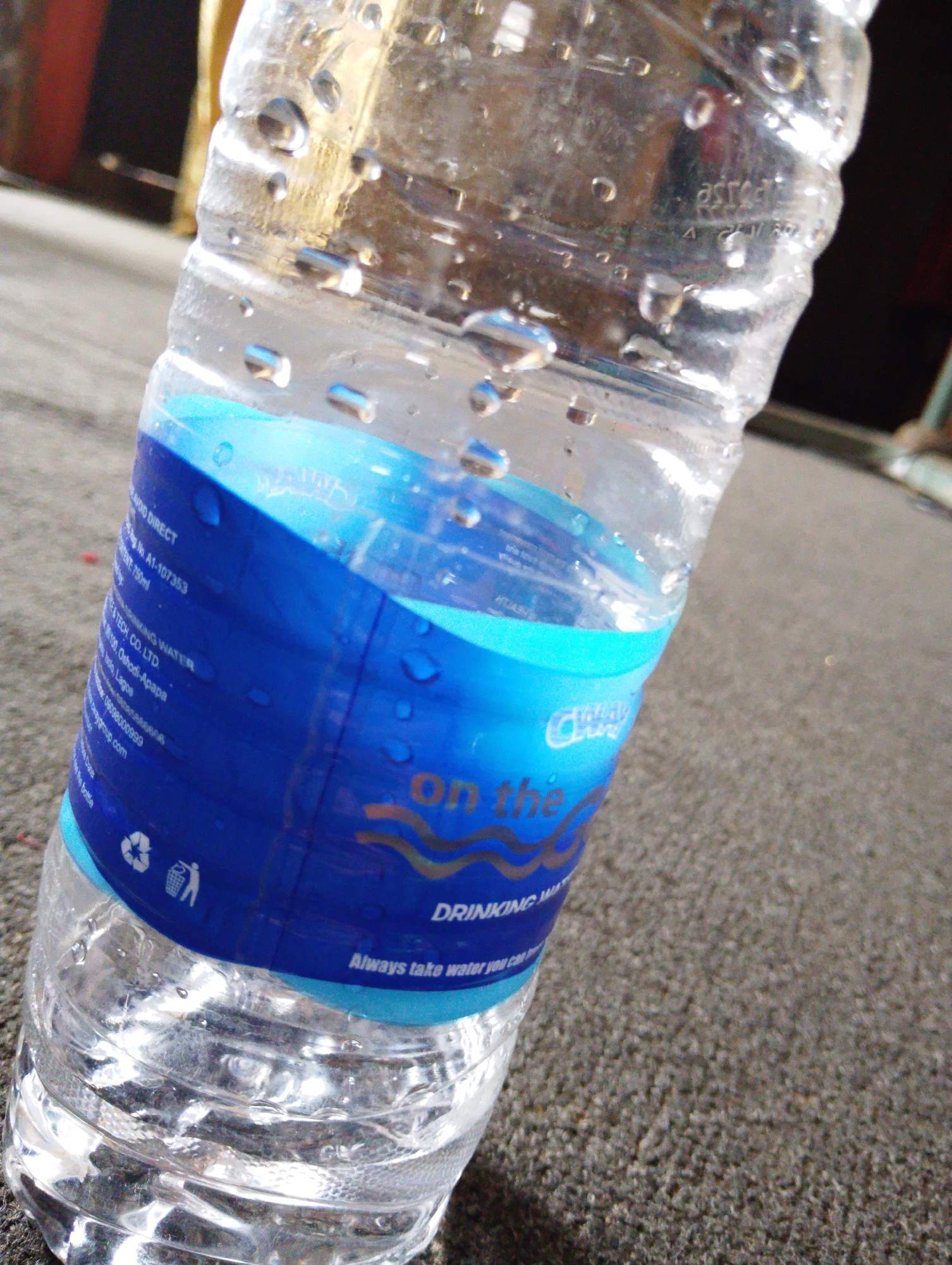 A close-up, eye-level shot focuses on a partially filled plastic water bottle. The bottle is covered in condensation droplets, and the label is predominantly blue and white with text. Part of the label reads "ON THE" in large, stylized font, with "DRINKING WATER" in smaller text below it. There are also recycling symbols visible on the label. The bottle rests on a textured, dark gray surface, possibly asphalt or coarse concrete. The background is blurred, showing hints of out-of-focus objects, including what appears to be a brown and yellow cylindrical object on the left and dark shapes in the upper right. The lighting suggests an outdoor setting, likely during daylight.