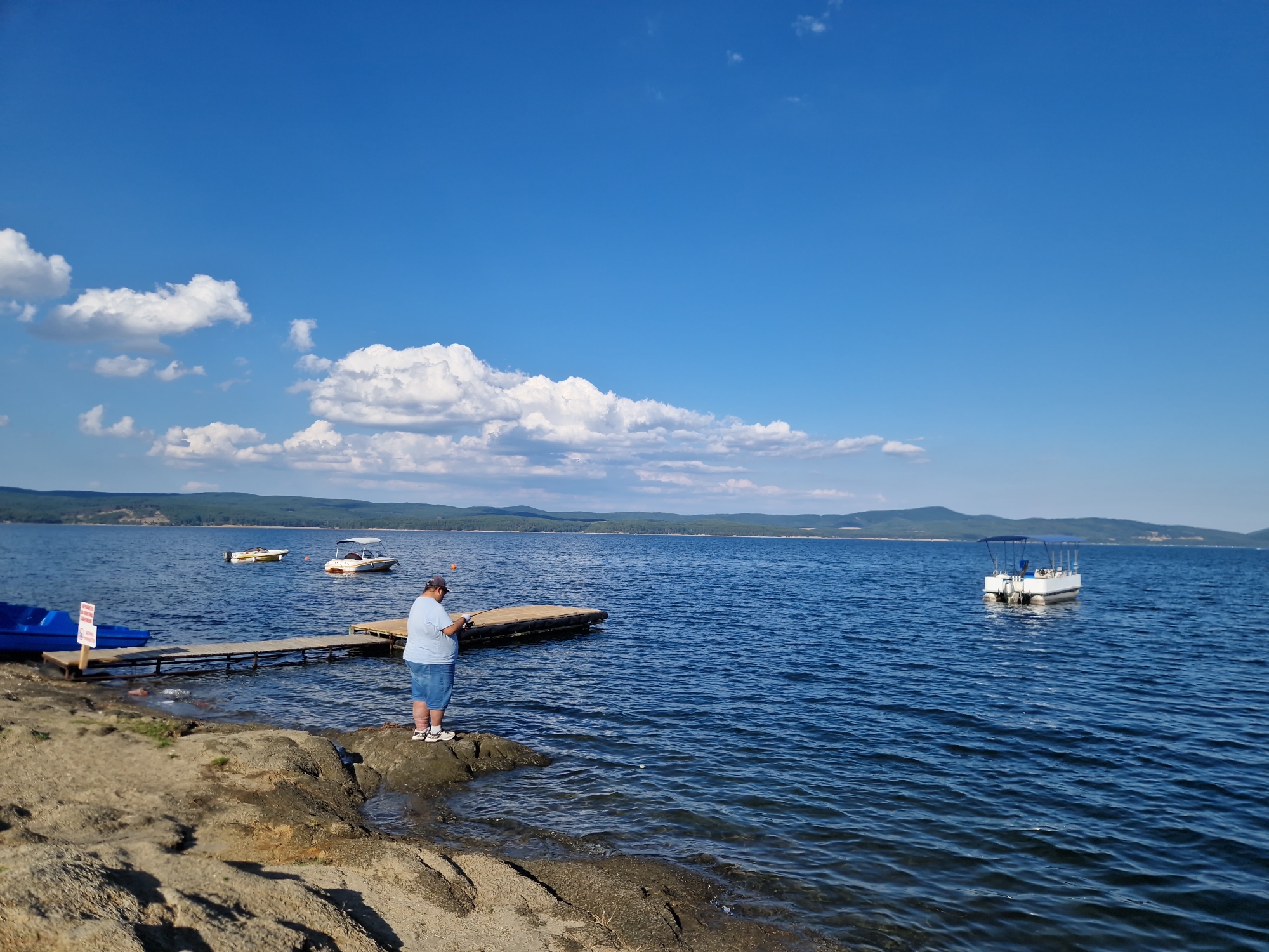 A man stands on a rocky shore, fishing in a wide, calm body of water under a bright blue sky with scattered white clouds. In the foreground, the shore is composed of large, tan-colored rocks and some patches of dry grass. To the left, a bright blue paddleboat is partially visible, pulled up onto the shore. A wooden pier extends into the water, with two small boats moored nearby. One is a yellow, flat-bottomed boat, and the other is a white boat with a canopy. Further out in the water, a larger white pontoon boat is anchored. In the background, rolling green hills covered in trees line the horizon, indicating a natural, possibly rural setting. The overall scene suggests a peaceful day at a lake or reservoir, likely near Dolni Pasarel, Bulgaria, given the context.