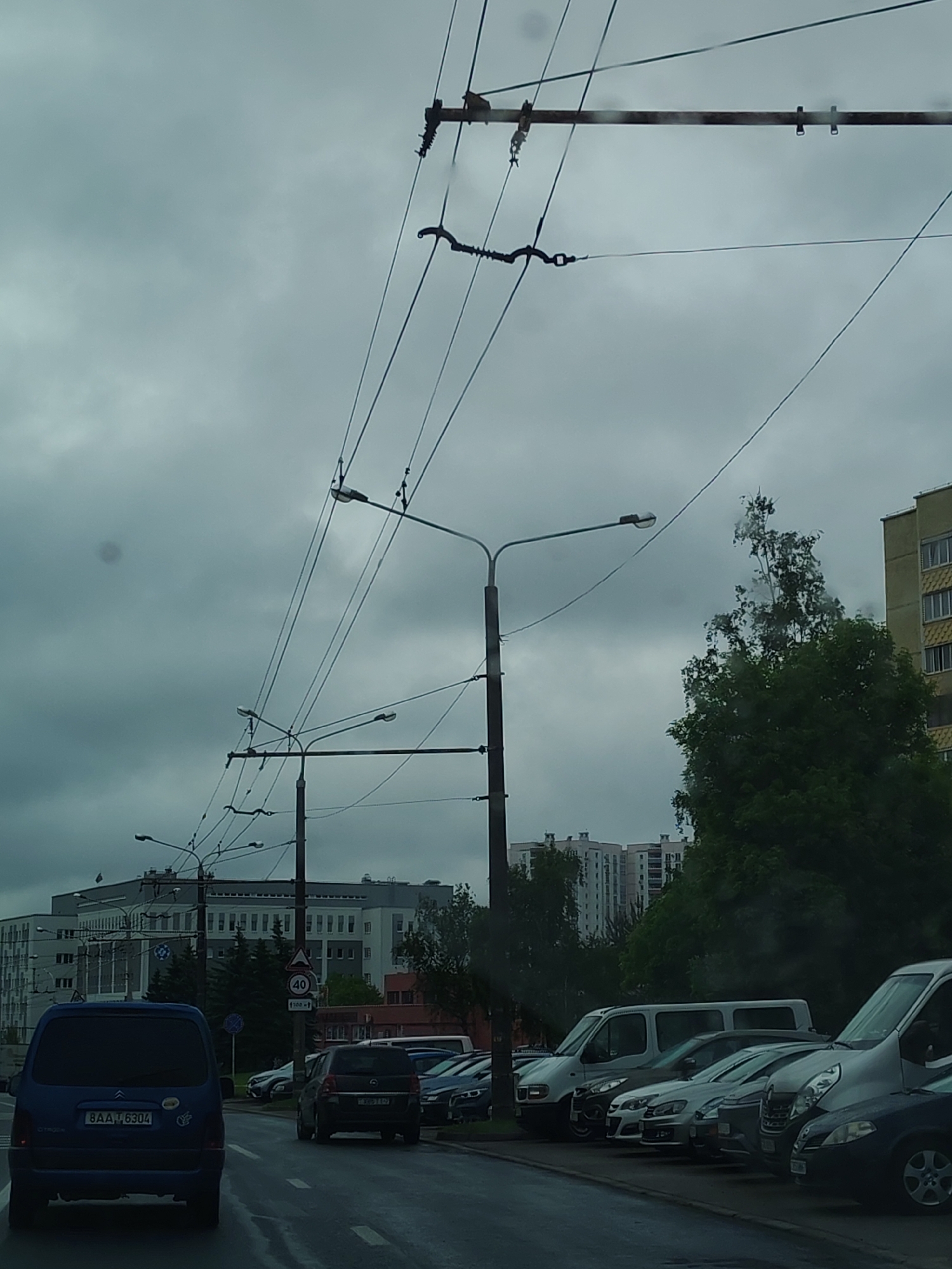 The image captures a street scene in an urban setting under a heavily overcast sky. Overhead, a complex network of tram or trolleybus power lines is visible, supported by black metal poles equipped with streetlights.
On the street, several vehicles are present. In the foreground, a dark blue Citroen Berlingo with license plate "BAA-76304" is positioned on the left side of the road, directly facing the camera. Further ahead and slightly to the right, a dark-colored Opel or similar vehicle is also moving along the road. A line of parked vehicles, predominantly vans and cars in shades of gray and silver, are parked along the right side of the street.
In the background, a mix of architectural styles is observed. A long, low-rise building with a repetitive window pattern is visible on the left. Adjacent to the parked vehicles and behind a cluster of trees, taller apartment buildings are located. A "40" speed limit sign (presumably kilometers per hour) is positioned on the street, indicating traffic regulations in the area.
