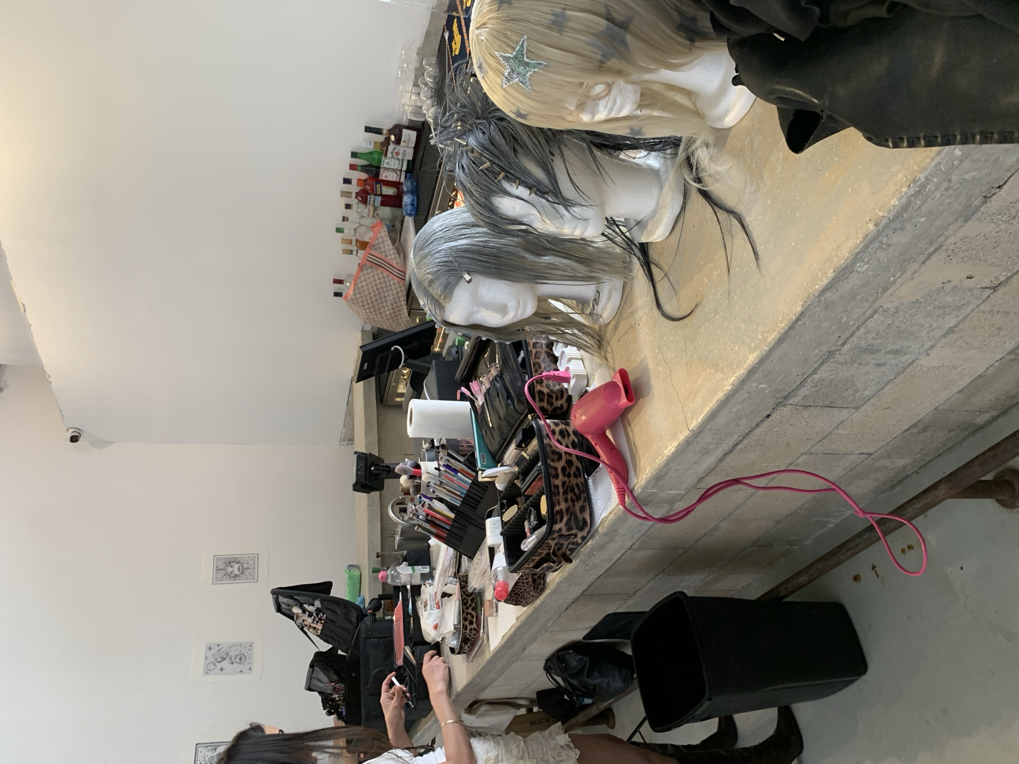 The image depicts a preparation area, possibly a makeup or hairstyling station, located indoors. Three mannequin heads with wigs are positioned on a counter covered with various cosmetic products, tools, and accessories. These include makeup brushes, palettes, and a pink hair dryer. The counter also holds a collection of liquor bottles. A woman is partially visible in the lower left corner of the frame, holding a small object in her hand. A black waste bin sits on the floor next to the counter. Two framed artworks hang on the white wall behind the counter, which is also equipped with a security camera. Based on the visual clues like the presence of Hebrew labels on some products and the overall architectural style, the location is likely in Tel-Aviv, Israel.