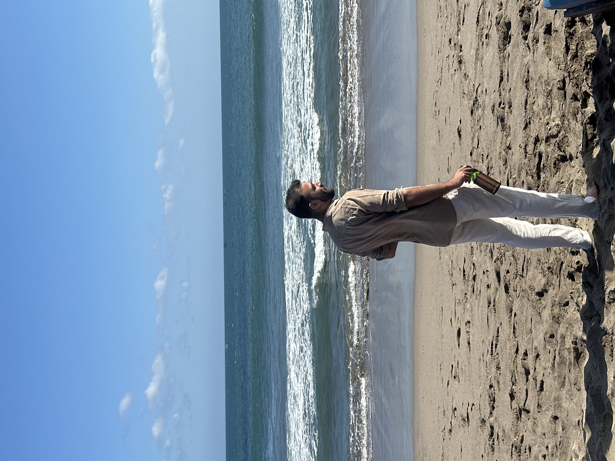 A man stands on a sandy beach facing away from the camera, looking out at the ocean. He is wearing a light brown button-down shirt and white pants. He holds a green and brown bottle in his right hand. The ocean has moderate waves with white foam. The sky is blue with scattered white clouds. The sand is light brown and has footprints and indentations. The location is the beach in North Kuta, Indonesia.