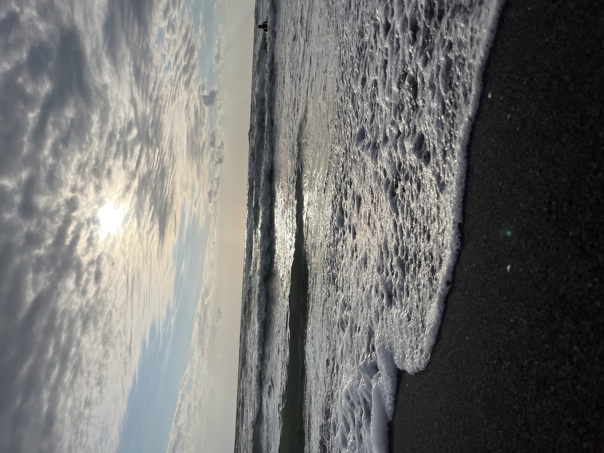A low-angle, vertical, eye-level shot captures a beach scene in Ureki, Georgia. The foreground is dominated by dark, wet sand, with a wave of foamy white water gently lapping at the shore. In the mid-ground, the sea extends outwards with successive waves rolling towards the viewer, reflecting the soft light from the sky. The water is a mix of dark blue and grey, with white crests on the waves. In the far distance, a single person is visible in the water, appearing as a small dark silhouette. The background features a cloudy sky with patches of sunlight breaking through, creating a diffused glow. The clouds are a mix of grey and white, with a hint of blue sky peeking through in the upper left corner. The overall atmosphere is serene and tranquil, with the sound of the waves implied.