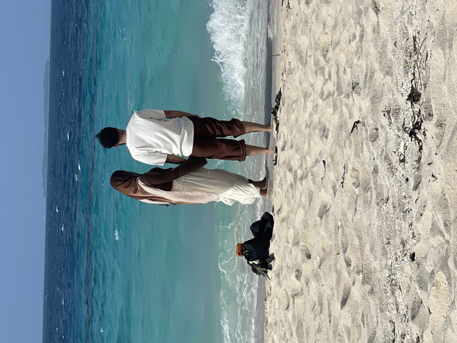 The image displays a beach scene in Zanzibar, Tanzania, under a clear sky. Two individuals stand with their backs to the viewer, facing a turquoise ocean, holding hands. The individual on the left wears a brown hijab, a brown long-sleeved top, and a light-colored long skirt. The individual on the right wears a white t-shirt and dark brown cropped pants. Both are barefoot, with their feet partially submerged in shallow water where small waves break. On the light-colored sandy beach in the foreground, a black backpack with an orange-cased smartphone propped on its side is visible. Scattered dark seaweed and small debris are present on the sand near the waterline. The ocean in the background transitions to a deeper blue, with a distant landmass visible on the horizon.