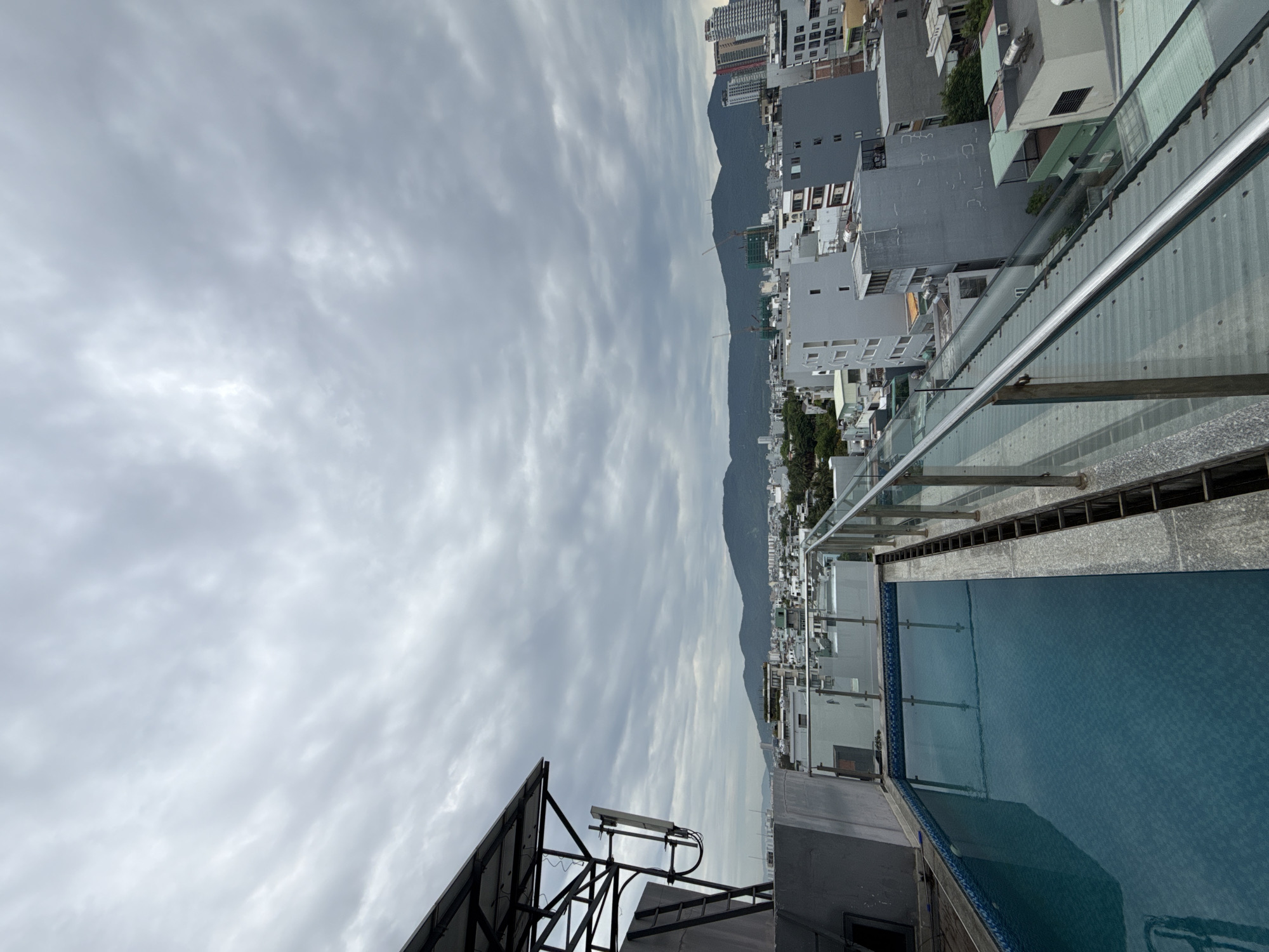 The image shows an elevated view of Đà Nẵng, Vietnam, under an overcast sky. The foreground includes a rooftop swimming pool with clear railing. In the distance, a mountain range is visible behind the city buildings. Cityscape includes both low-rise buildings and high-rise buildings, indicating a mix of residential and commercial development.