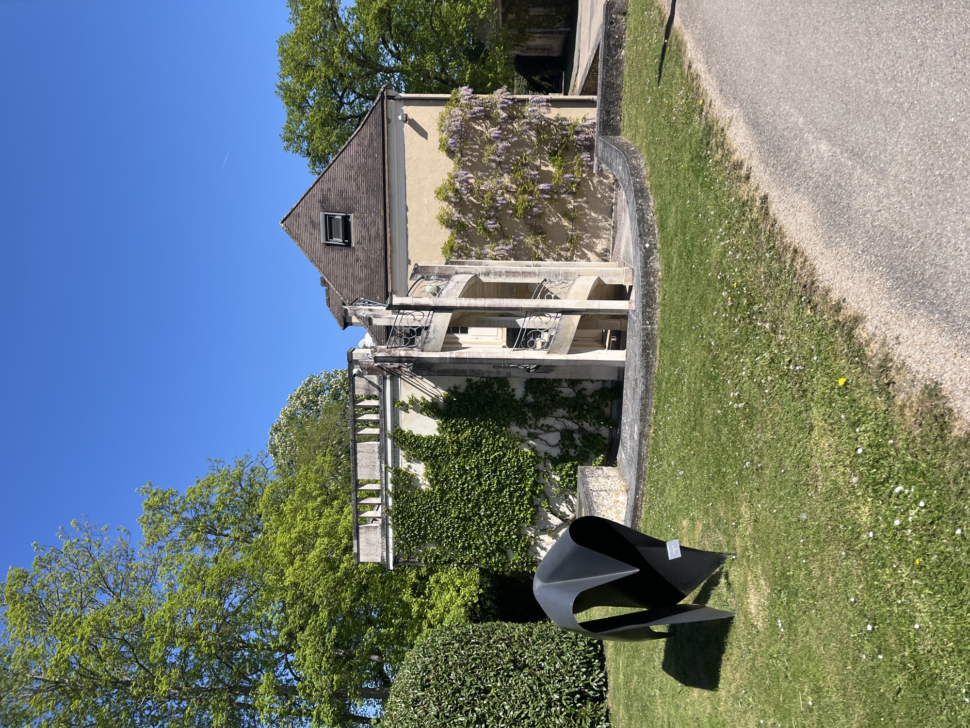 The image shows a building with a light yellow facade, covered in parts with climbing ivy and wisteria vines. The building has a brown tiled roof with a small window. The building has a balconied extension. In the foreground is a modern black sculpture, positioned on a green lawn dotted with daisies. To the right of the sculpture is a paved pathway. Green trees are also present. The location is Saint-Rémy-lès-Chevreuse, France.