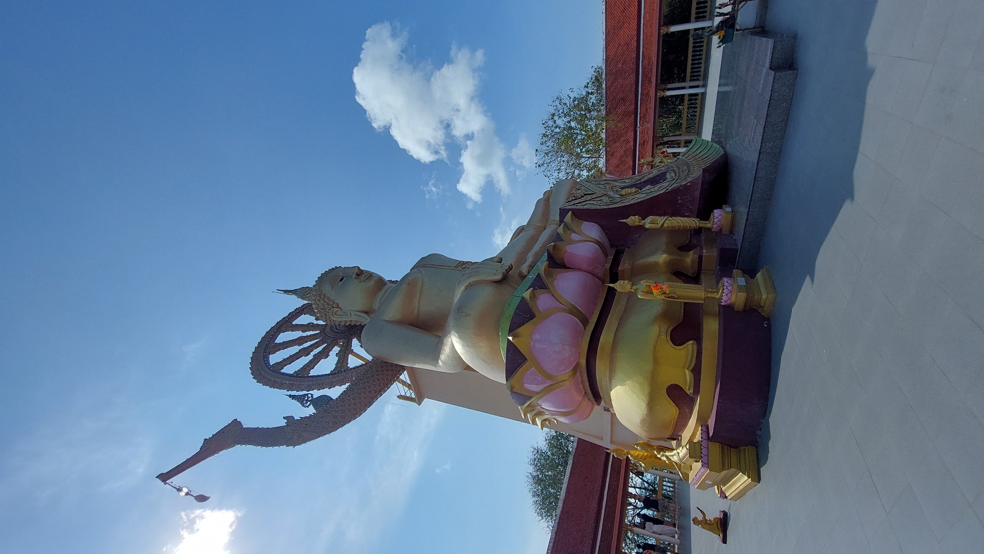 The image captures a majestic golden reclining Buddha statue under a bright blue sky, located in Baan Plai Laem, Thailand. The large statue lies horizontally, with its head resting on its right arm, facing the left side of the frame. Behind the Buddha's head is an elaborate, golden, spoked wheel, likely representing the Dharmachakra or Wheel of Dharma.
The Buddha rests on a richly decorated pedestal featuring vibrant pink lotus flower motifs and intricate golden patterns. Along the base of this pedestal, several smaller golden standing Buddha statues are visible, some adorned with small orange floral garlands.
In the background, the sky is a clear, brilliant blue with a few scattered white cumulus clouds. To the right of the main statue, part of a temple building with a distinctive reddish-orange tiled roof and dark architectural elements is visible, nestled among green trees. The ground in the foreground is a light grey paved surface, with distinct shadows cast by the statue and surrounding structures, indicating strong overhead sunlight during the day. No visible text is present in the image. The scene conveys a sense of grandeur and spiritual serenity typical of a Buddhist temple setting.