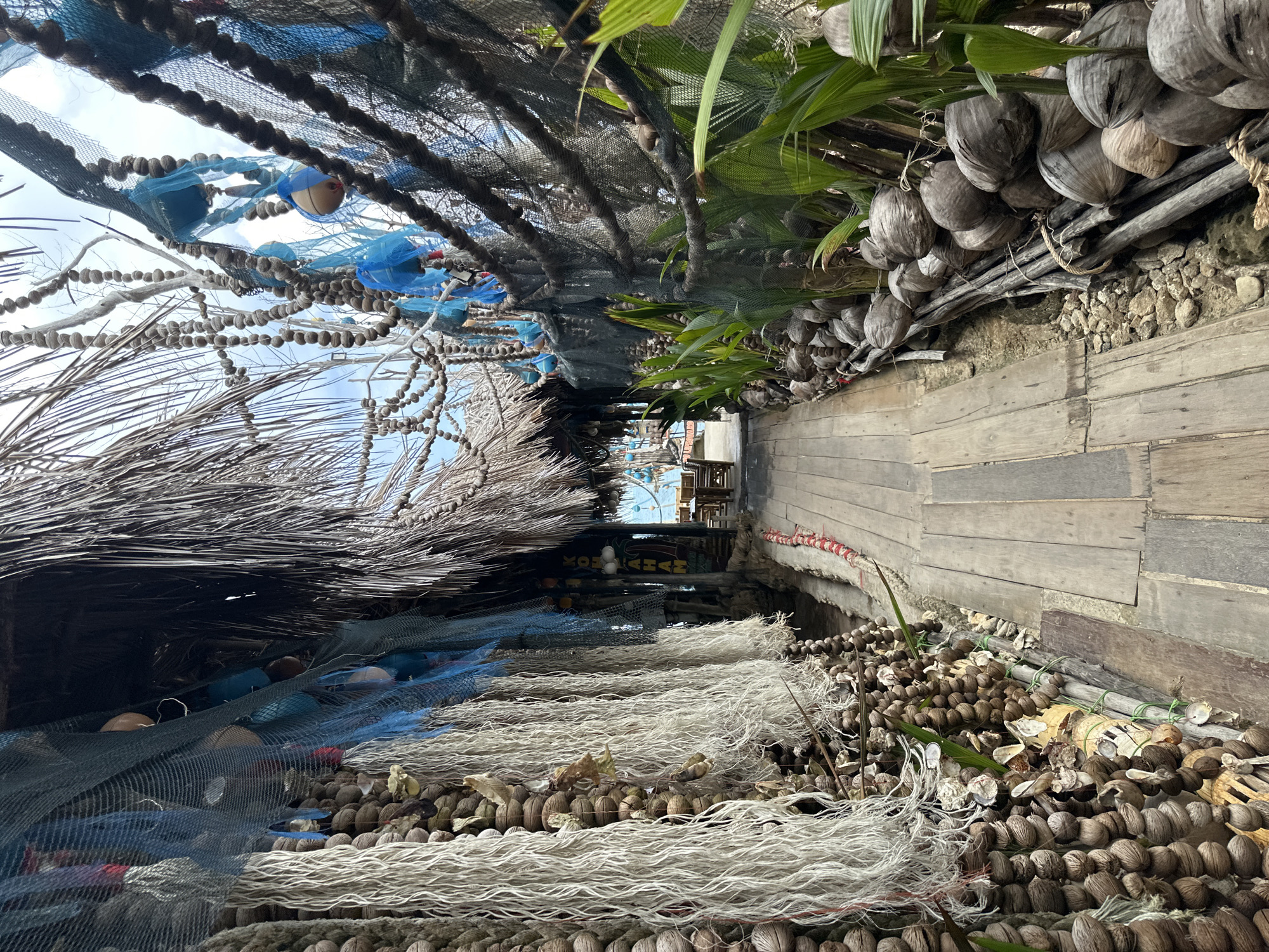This image depicts a narrow wooden walkway leading through an outdoor establishment on Koh Phangan, Thailand. The walkway is flanked by displays of dried coconuts and shells, as well as various forms of netting and ropes, some adorned with blue mesh bags and spherical buoys. These decorative elements hang from structures covered in thatch and palm fronds, suggesting a tropical, beachside setting. In the background, tables and chairs are visible, hinting at a seating area or restaurant. The overall impression is one of a rustic, island-themed venue.