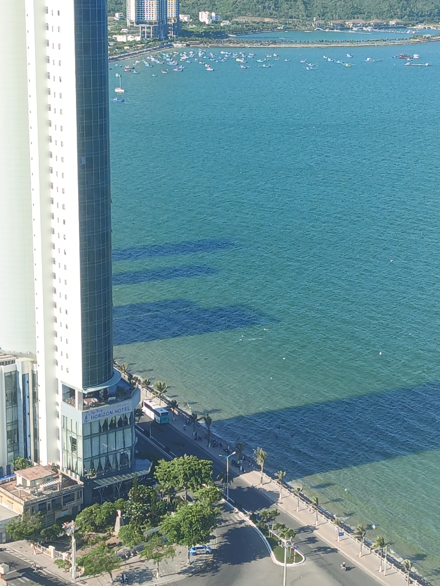 The image shows a high-angle view of a coastal city, likely in Bac Nha Trang Ward, Vietnam. A large, modern white building with a cylindrical glass section is prominent on the left. This building appears to be the "HORIZON HOTEL," as indicated by signage on the glass section. The hotel has multiple floors, with visible windows and balconies. Adjacent to the hotel, there are lower-rise buildings and green trees.
To the right of the buildings is a body of blue water, possibly the sea, dotted with numerous small boats. The water is relatively calm, with some shadows cast upon it from the buildings and possibly clouds. A paved road runs along the coastline in front of the hotel, with palm trees lining the sidewalk. Several vehicles, including a bus and motorcycles, are visible on the road and sidewalk. Pedestrians can also be seen walking along the sidewalk. In the background, there are more buildings and hills. The overall scene depicts a vibrant urban coastal environment.