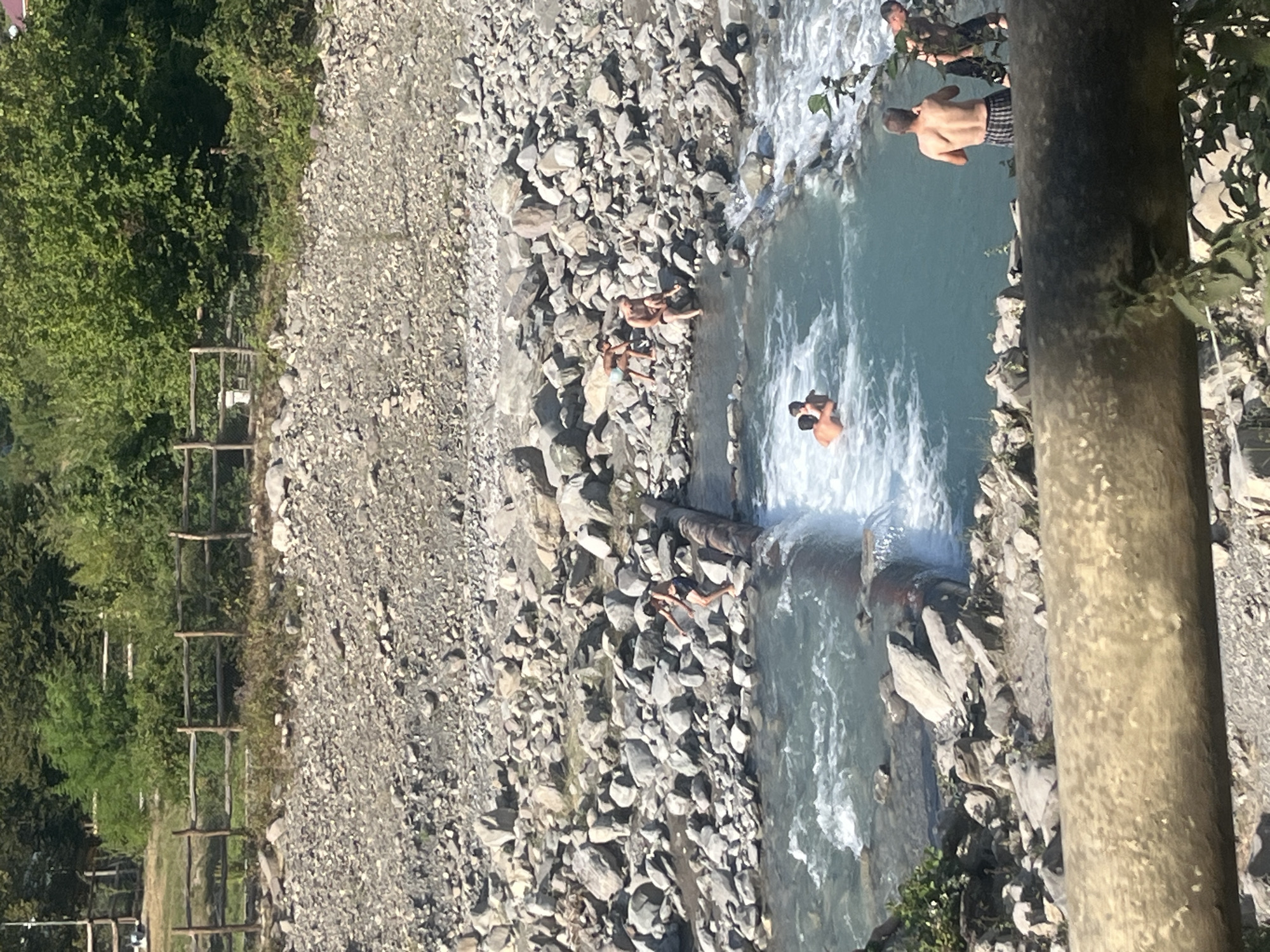 In Ismailli District, Azerbaijan, a group of people are enjoying a sunny day at a shallow, rocky river. The river has a small dam made of logs, creating a waterfall. Several individuals are in the water, some swimming and playing, while others sit on the rocks along the riverbank. The surrounding environment is characterized by dense green foliage and a rocky, uneven terrain. The scene depicts a recreational area where people are engaging in water activities amidst a natural, rugged landscape.