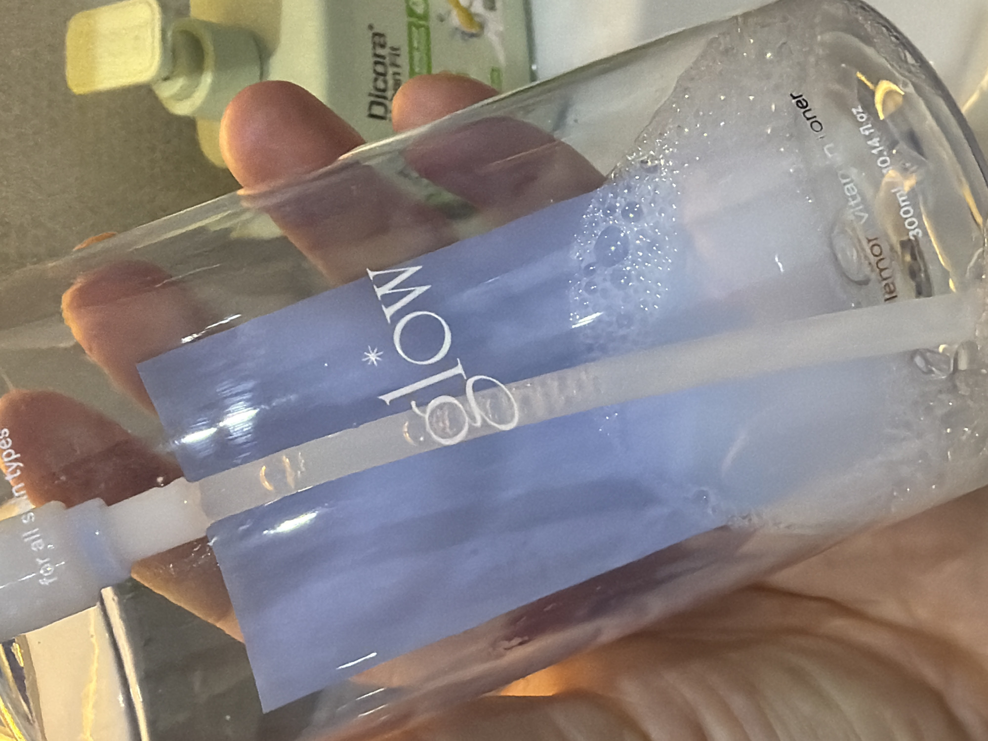 A close-up shot depicts a clear plastic bottle held in a person's hand. The bottle's label features the word "GLOW" in large, stylized letters. The bottle contains a frothy liquid. Text on the bottle indicates it is a "Lemnor Vitamin Toner" with a volume of "300ml/10.14 fl oz". Another text reads "for all skin types". The background features other bottles and a textured surface. The location is not specified.
