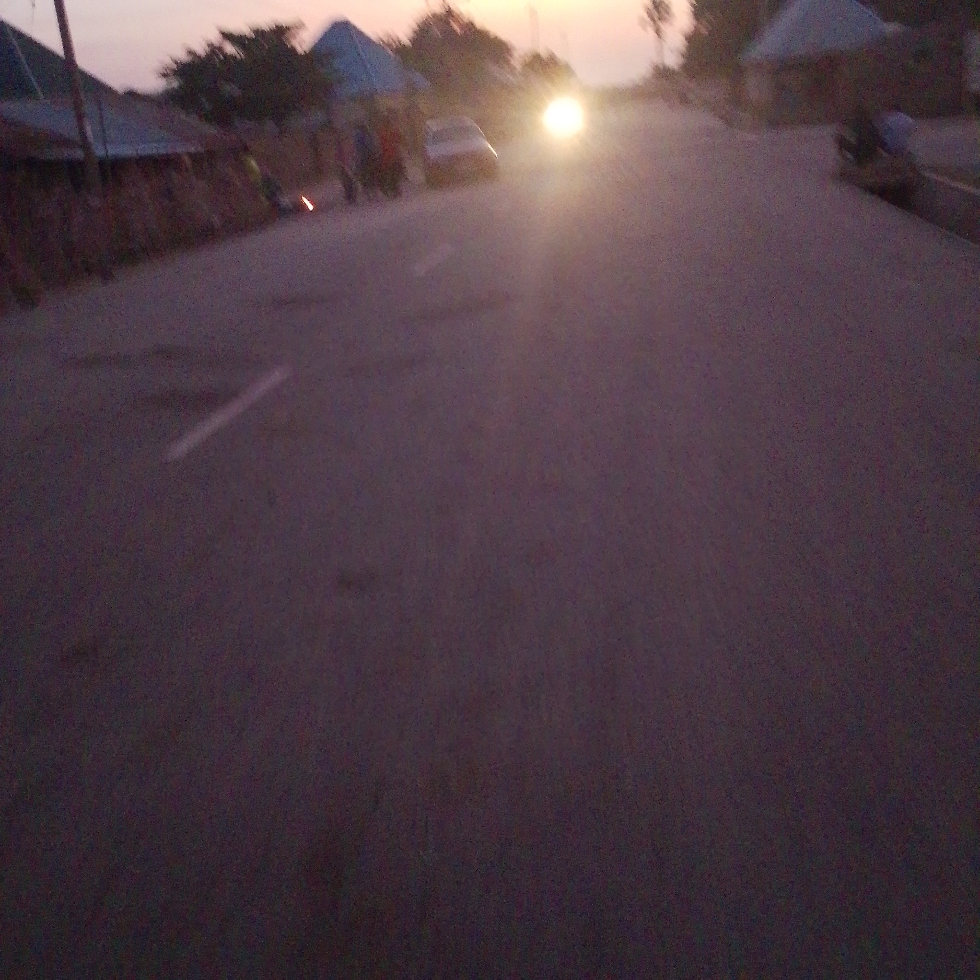 The image depicts a street scene at dusk in Wuro Allahamdu, Nigeria. A vehicle with its headlights on is prominent in the center of the frame, creating a bright glare. Several figures, appearing to be pedestrians, are visible on the left side of the road, some clustered together. To the left, there are structures that resemble buildings or market stalls with conical roofs. The road surface is asphalt, and a white line marking the lane is faintly visible. The overall lighting suggests it is either sunrise or sunset, with the sky showing a pale gradient. The environment appears to be a settlement or village area with some vegetation present in the background.