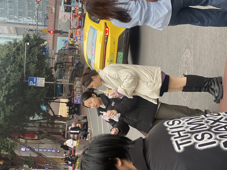 The image depicts an urban street scene in Yuzhong District, China, under overcast daylight conditions. In the mid-ground, two East Asian women stand on the edge of an asphalt road. The woman on the left, wearing a black puffer jacket over a white top and olive-green pants, holds a clear plastic cup with a straw near her mouth. The woman on the right, dressed in an off-white puffer jacket over a purple top, black shorts, and knee-high black boots, holds a similar clear plastic cup with a straw. Both have dark hair tied back. To their immediate right, the rear of a yellow new energy vehicle taxi, displaying the license plate '渝BTO297' and a blue sticker on its back window, is stationary in a lane of traffic beside a visible red traffic light.
In the foreground, partial views of two additional individuals are present: a person on the far left wearing a black hoodie with white text "THIS IS MY MAX" (partially visible), and a person on the far right wearing a light blue shirt and blue jeans.
The background features a multi-lane road with various vehicles, including a brown SUV, a large tan bus marked "BA17506," and other cars in different lanes. A blue parking sign with a white 'P' and Chinese text is prominently displayed. The left side of the street includes a sidewalk lined with green-leafed trees, planters containing yellow flowers, and commercial buildings. One building sign clearly reads "33轻奢酒店" (33 Light Luxury Hotel) in Chinese. Modern high-rise buildings are visible in the distant background, completing the urban environment.