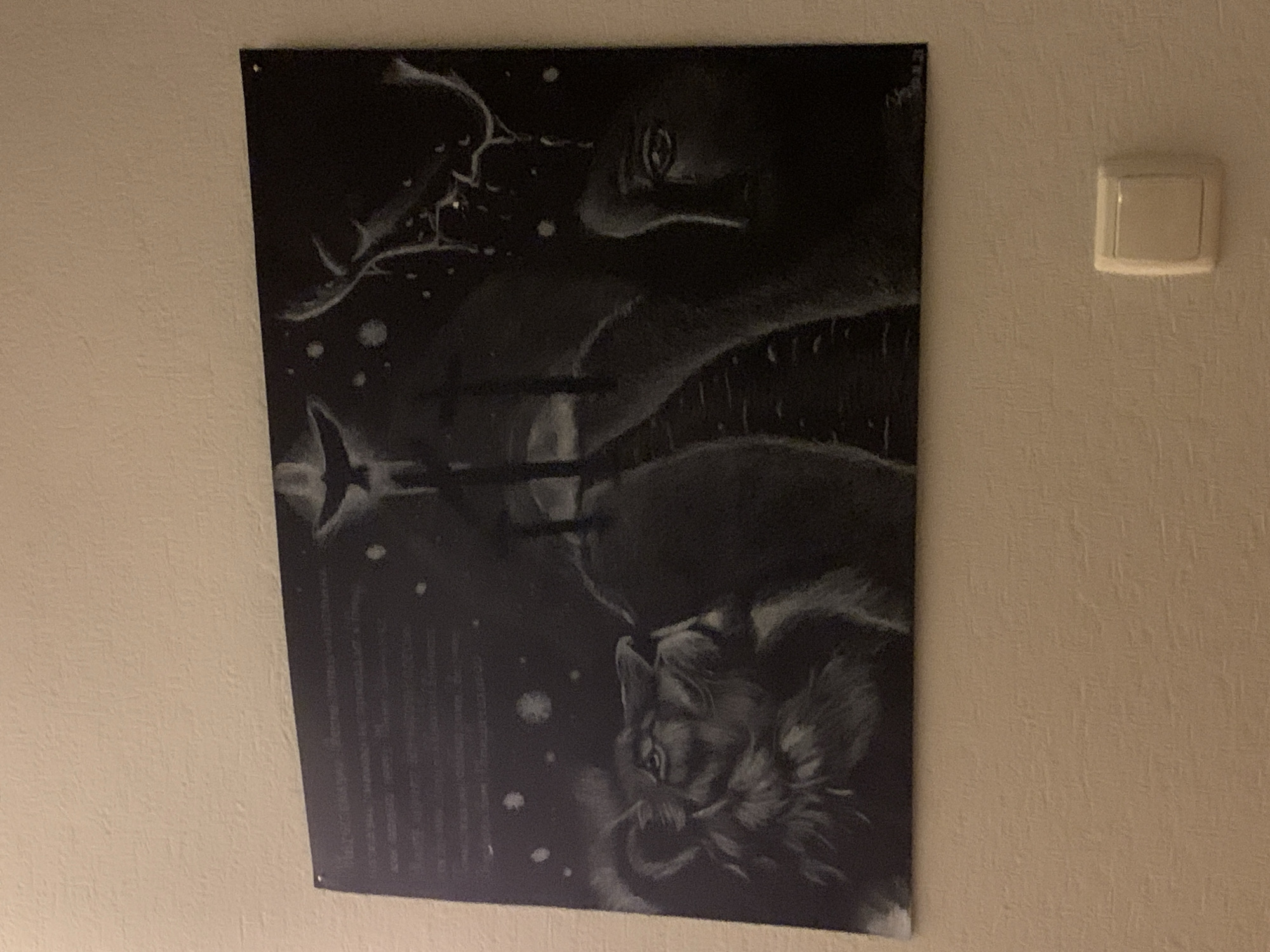 The image shows a black and white painting on a textured, beige wall. The painting is abstract and features a lion's head on the left, looking upwards. In the center, there are three figures with what appear to be halos, standing on a dark, indistinct path. Above them, a bird is silhouetted against a starry sky. On the right side of the painting, there is a profile of a human face, seemingly emerging from a dark, stormy background with lightning. The painting is attached to the wall with what appear to be two small pins or screws at the top. Below the painting, there is a light switch with a white cover. The overall scene appears to be an indoor setting, likely a room in a residential or public building. There is no discernible information in the image that would place it specifically within the Чыжэвіцкі сельскі Савет, Belarus.