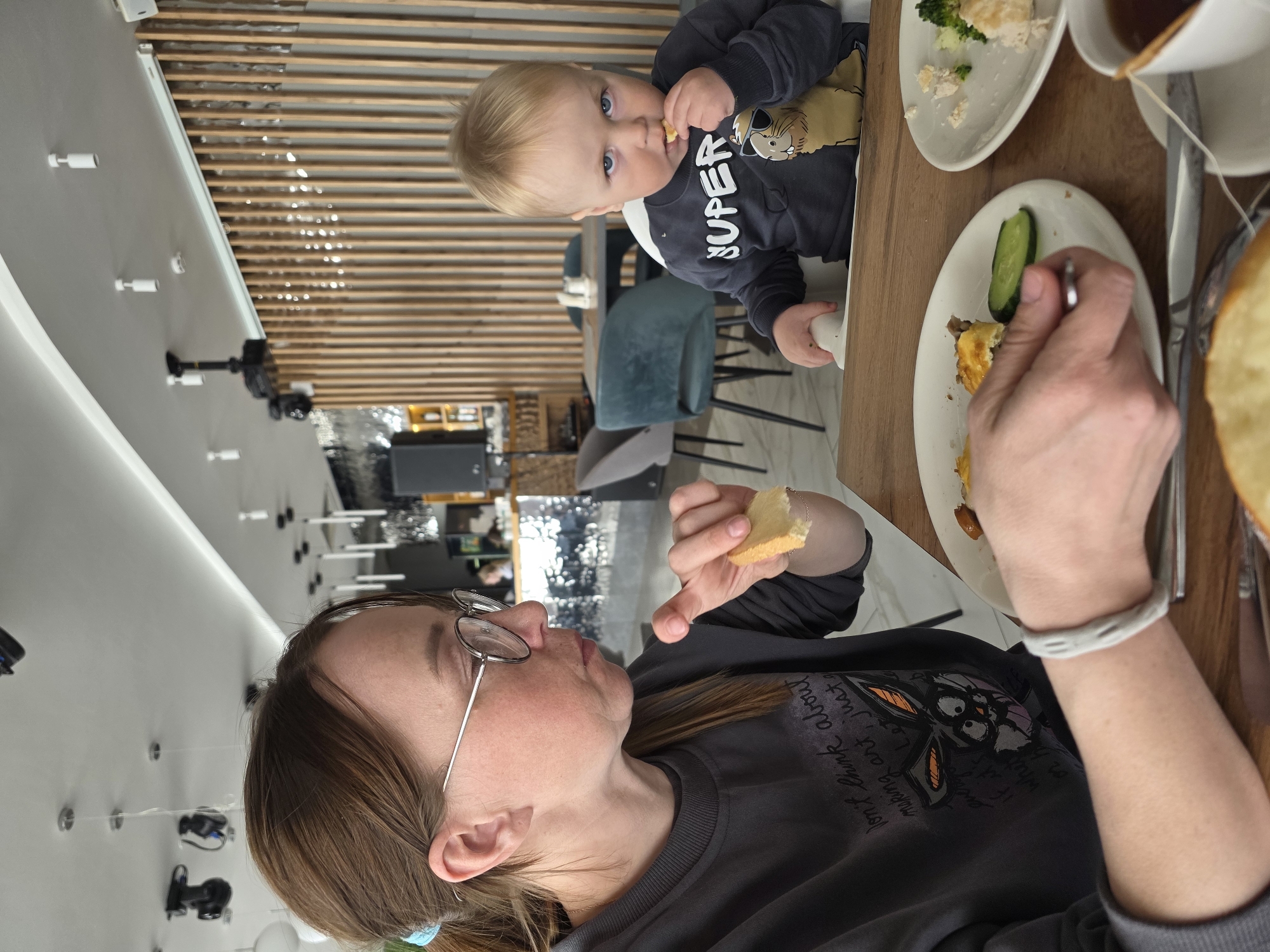 The image captures a scene in what appears to be a restaurant in Votkinsk, Russia. A woman is seated at a wooden table, wearing glasses and a dark gray sweatshirt with a graphic print. She is holding a piece of bread in her left hand and using a fork to eat from a white plate containing an omelet and cucumber. A bracelet is visible on her right wrist. To her left, a young child is sitting in a high chair, wearing a gray long-sleeve shirt with the word "SUPER" printed on it. The child is holding a piece of food and looking towards the camera. The table also has other plates with food, including one with broccoli and chicken. A cup with tea can also be seen. The background features a wall with a horizontal slat design and a view into the restaurant, including chairs and lighting fixtures.